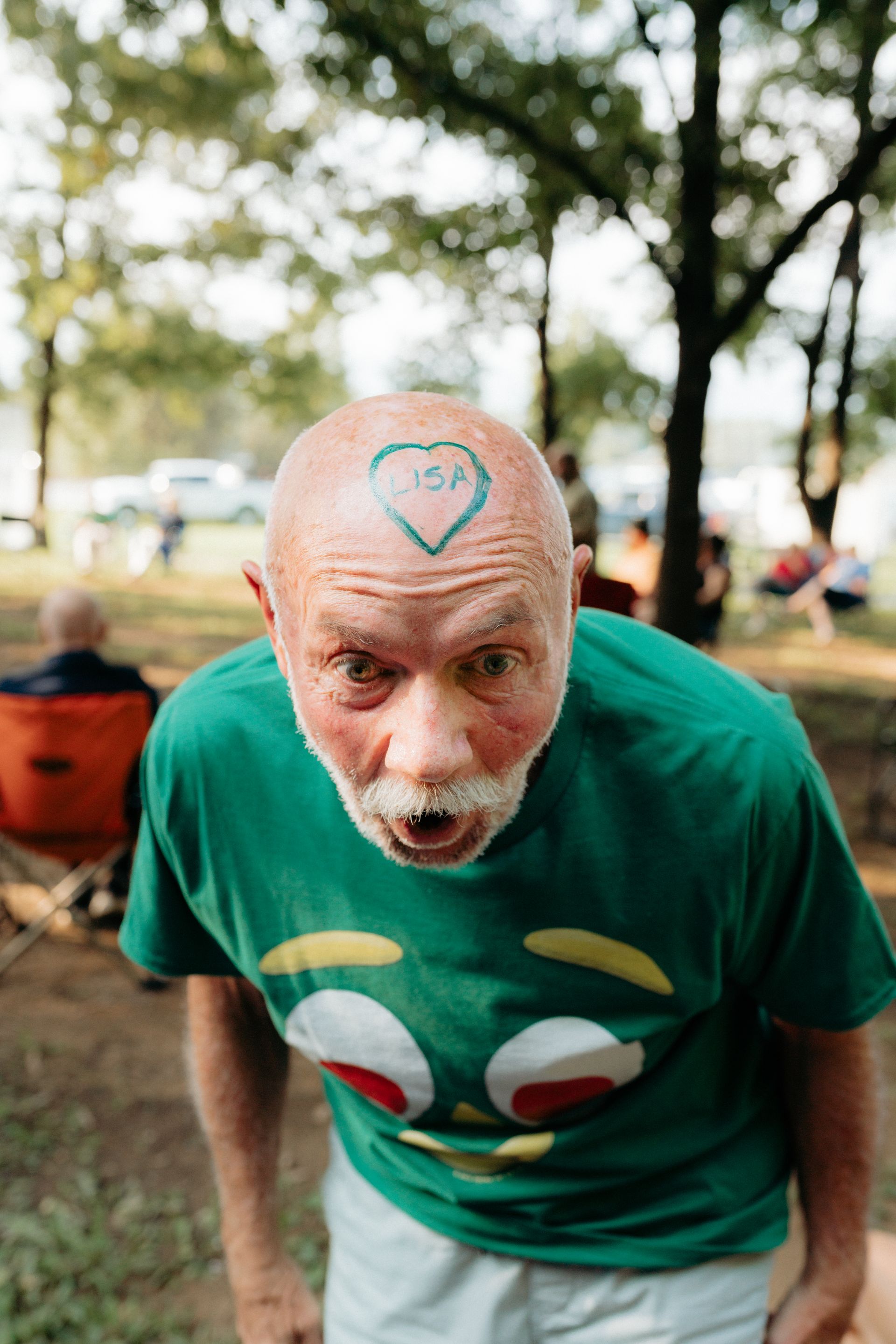 Bald man with a green shirt and heart tattoo on his head makes a surprised face outdoors.