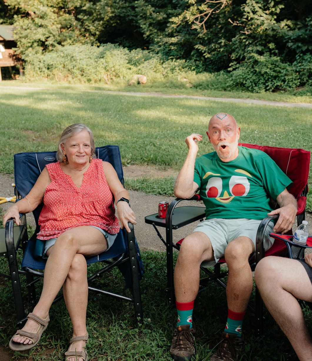 Two people sitting in camping chairs outdoors; woman in red, man in green shirt, both relaxed expressions.
