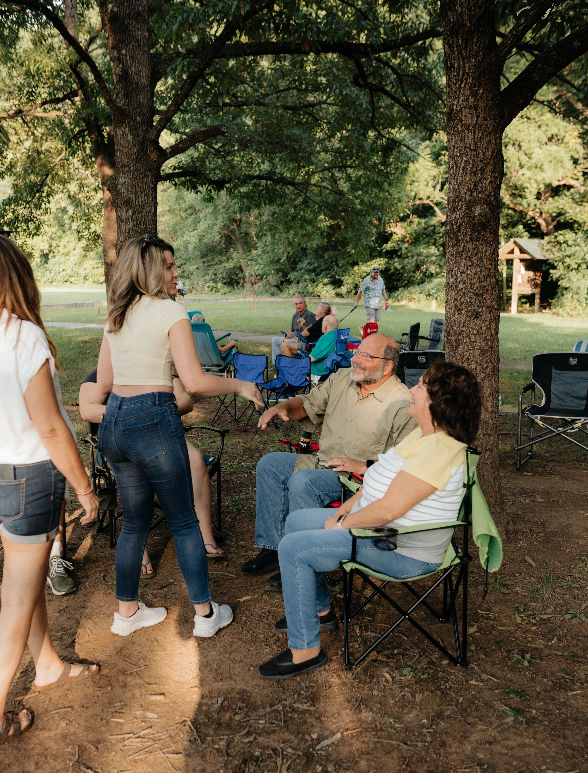 People gathered outdoors, under trees. A man and woman sit in chairs; others stand nearby.