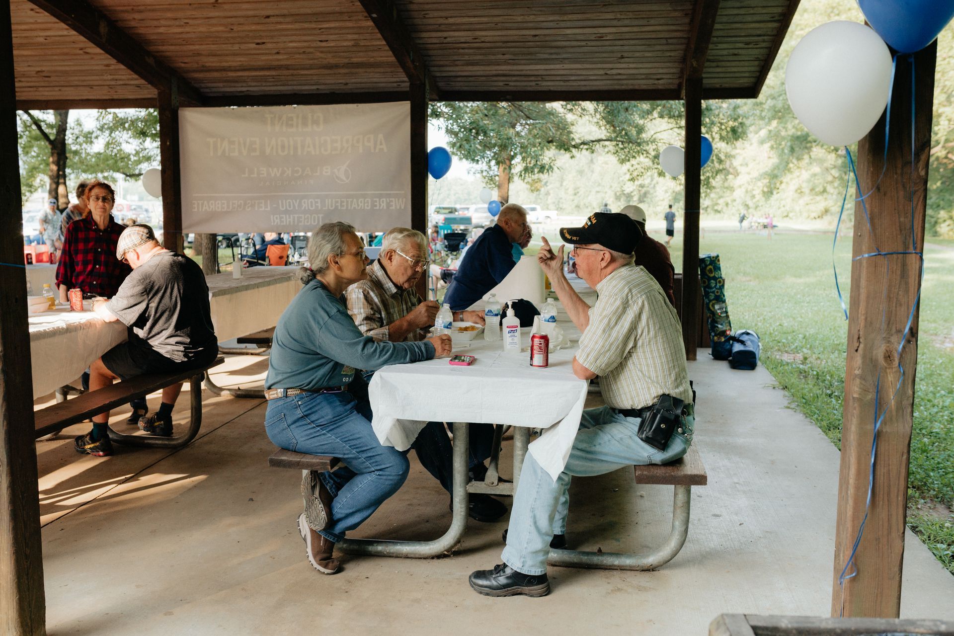 People eating and talking at a picnic table under a pavilion, outdoors. Balloons and a banner decorate the area.