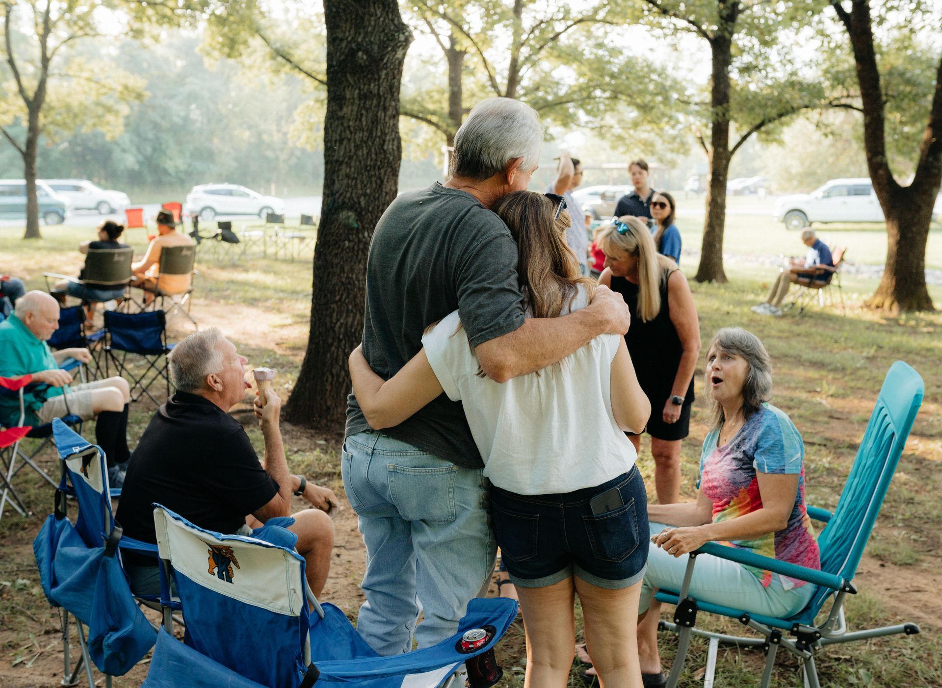 People embrace at outdoor gathering in park; other attendees in chairs and cars in background.