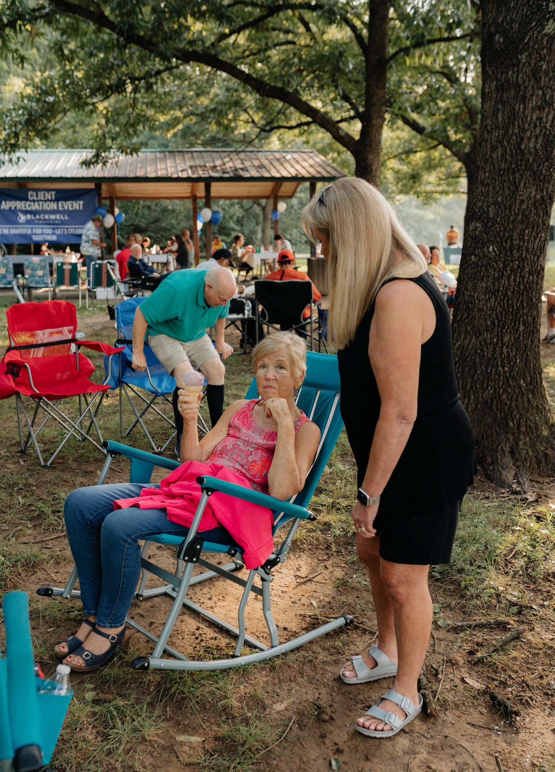 Three women at an outdoor gathering; one in a rocking chair, another standing and talking, the third behind them, with a pavilion in the background.