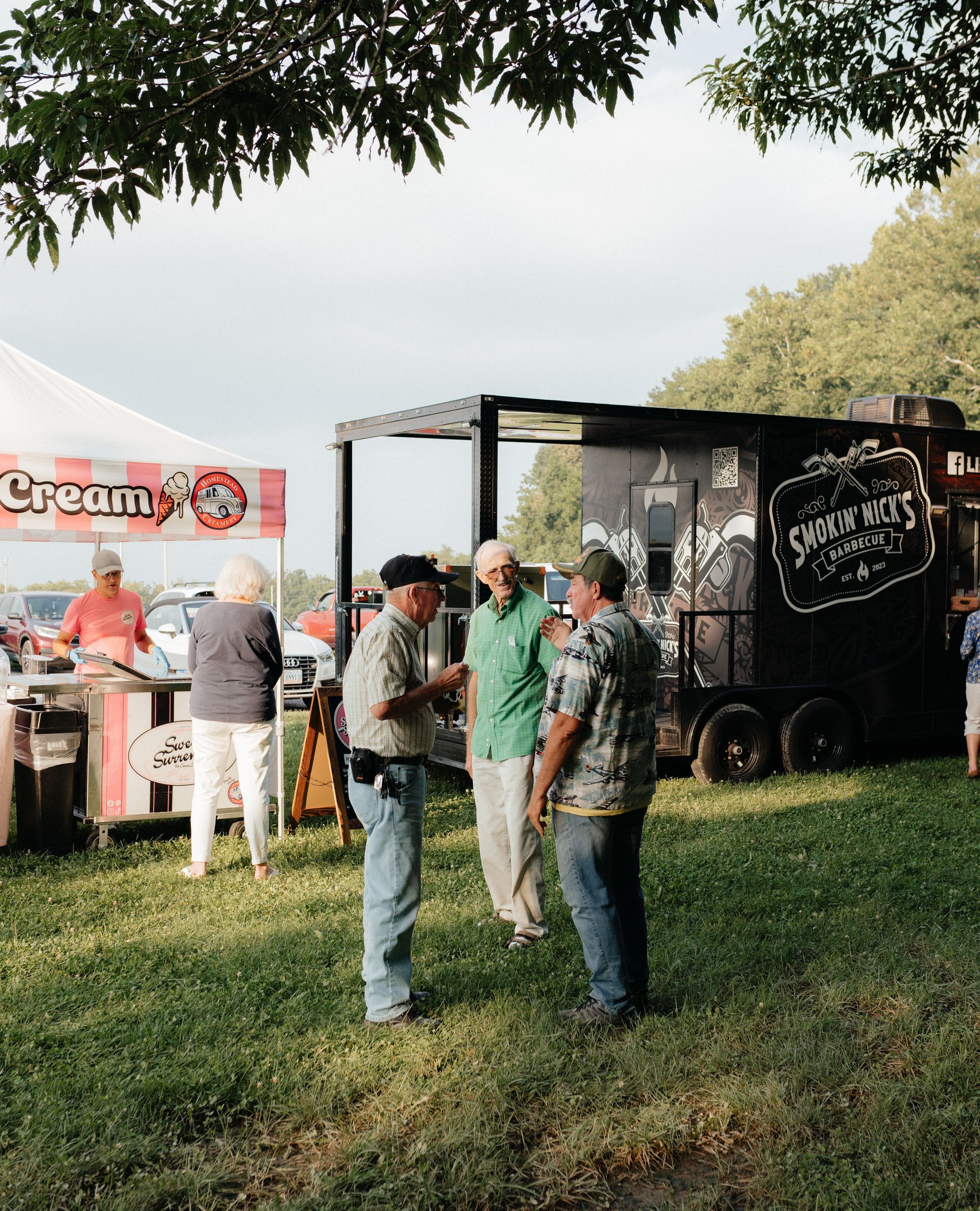 People at a food stand, socializing. Ice cream stand with a black food truck on grass, under trees.