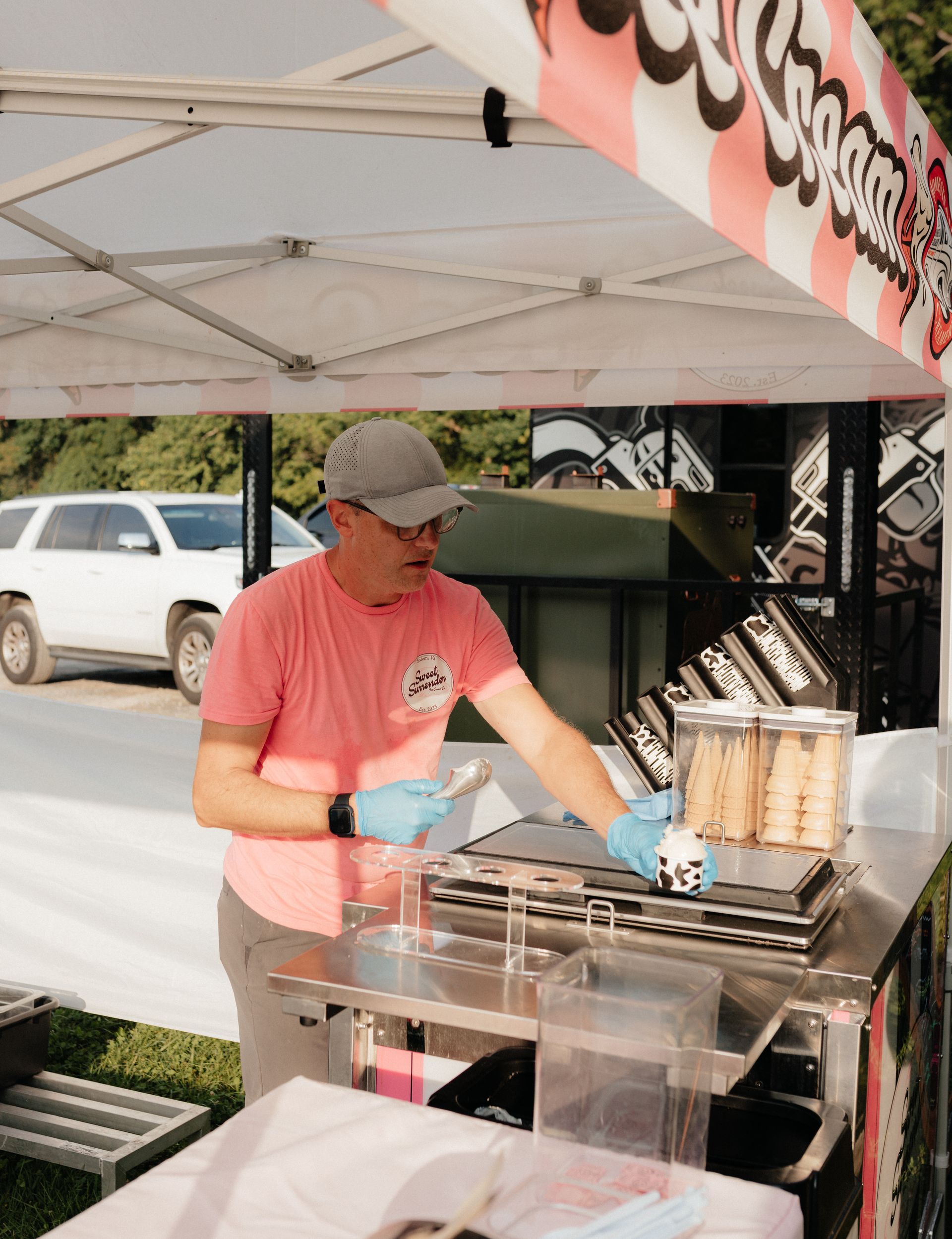 Man in pink shirt serving ice cream from a stand with pink and white awning. He wears gloves and a cap.