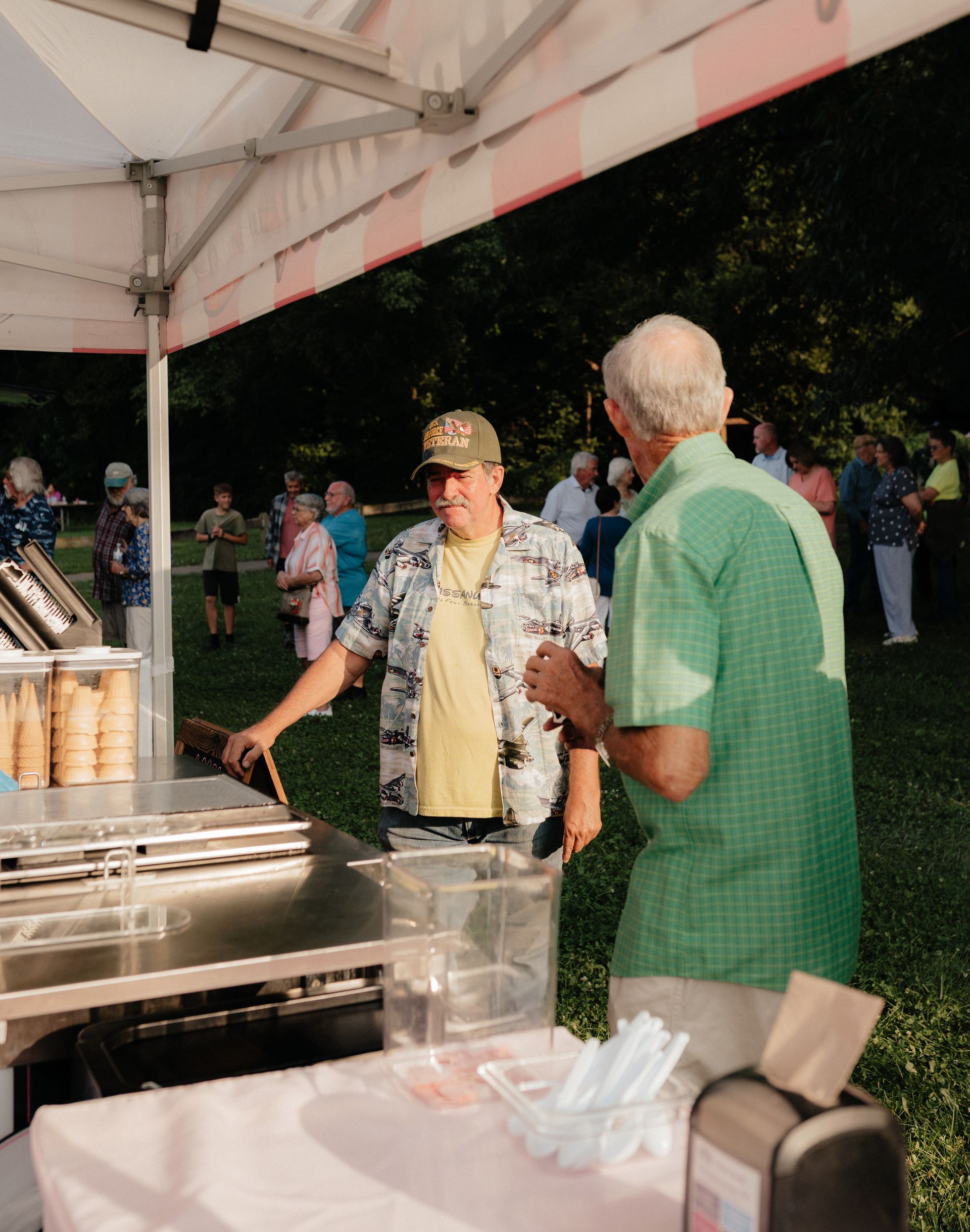 Two men at an ice cream stand. Man in green shirt talks to vendor under a tent.  A crowd of people in background.