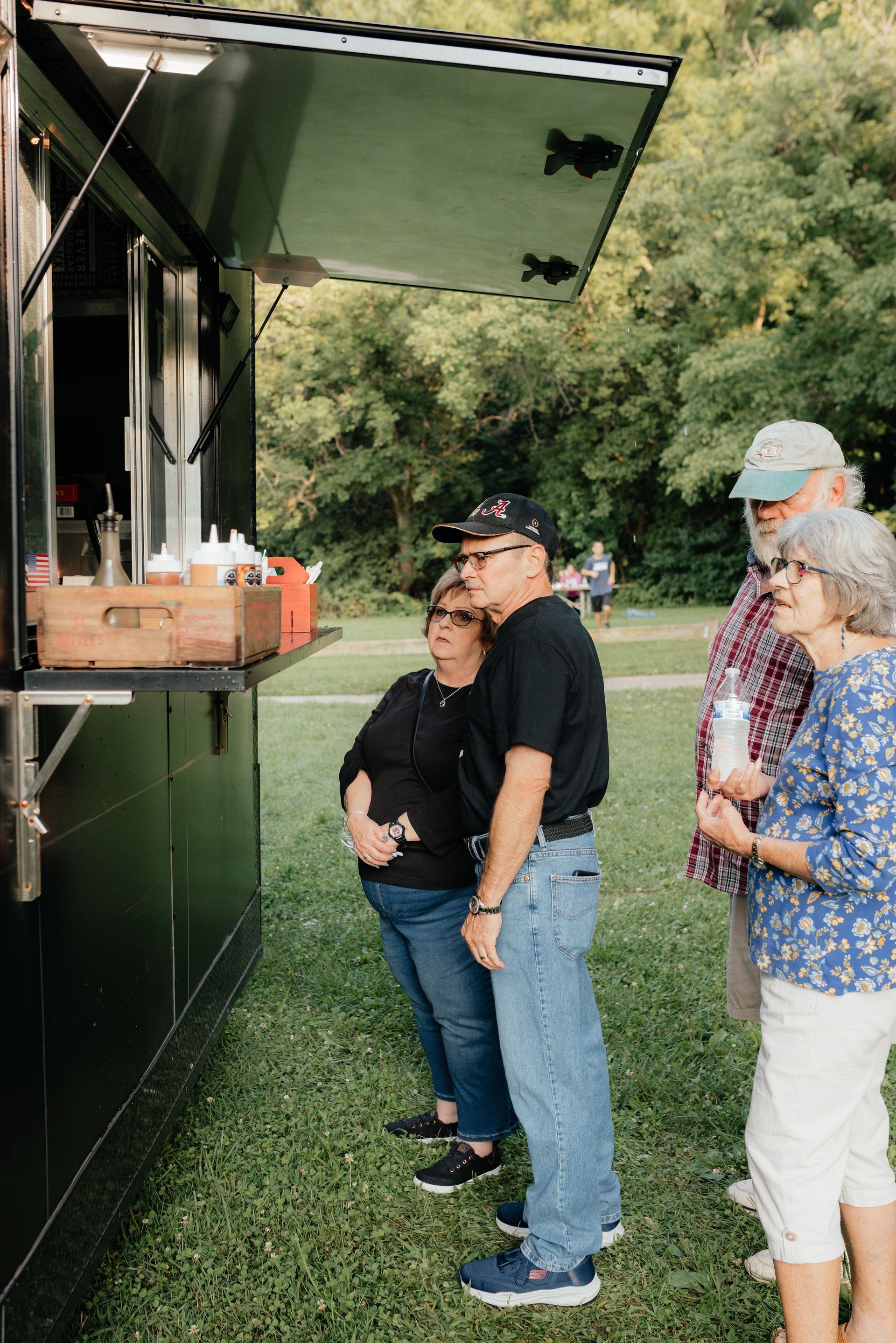 People waiting in line at a black food truck in a park setting.