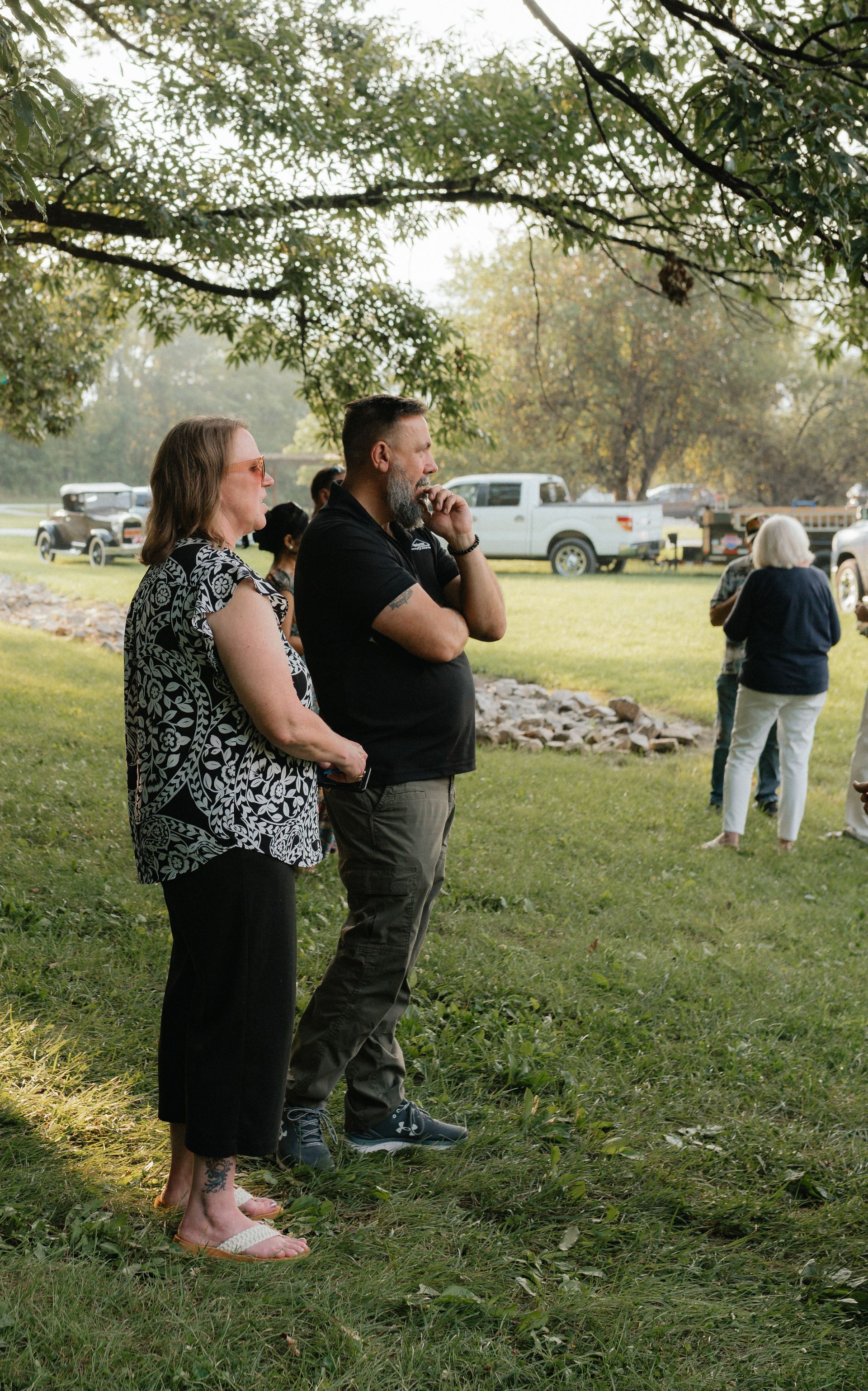 Woman and man standing outdoors, looking ahead. Man has beard. People and vehicles in background.