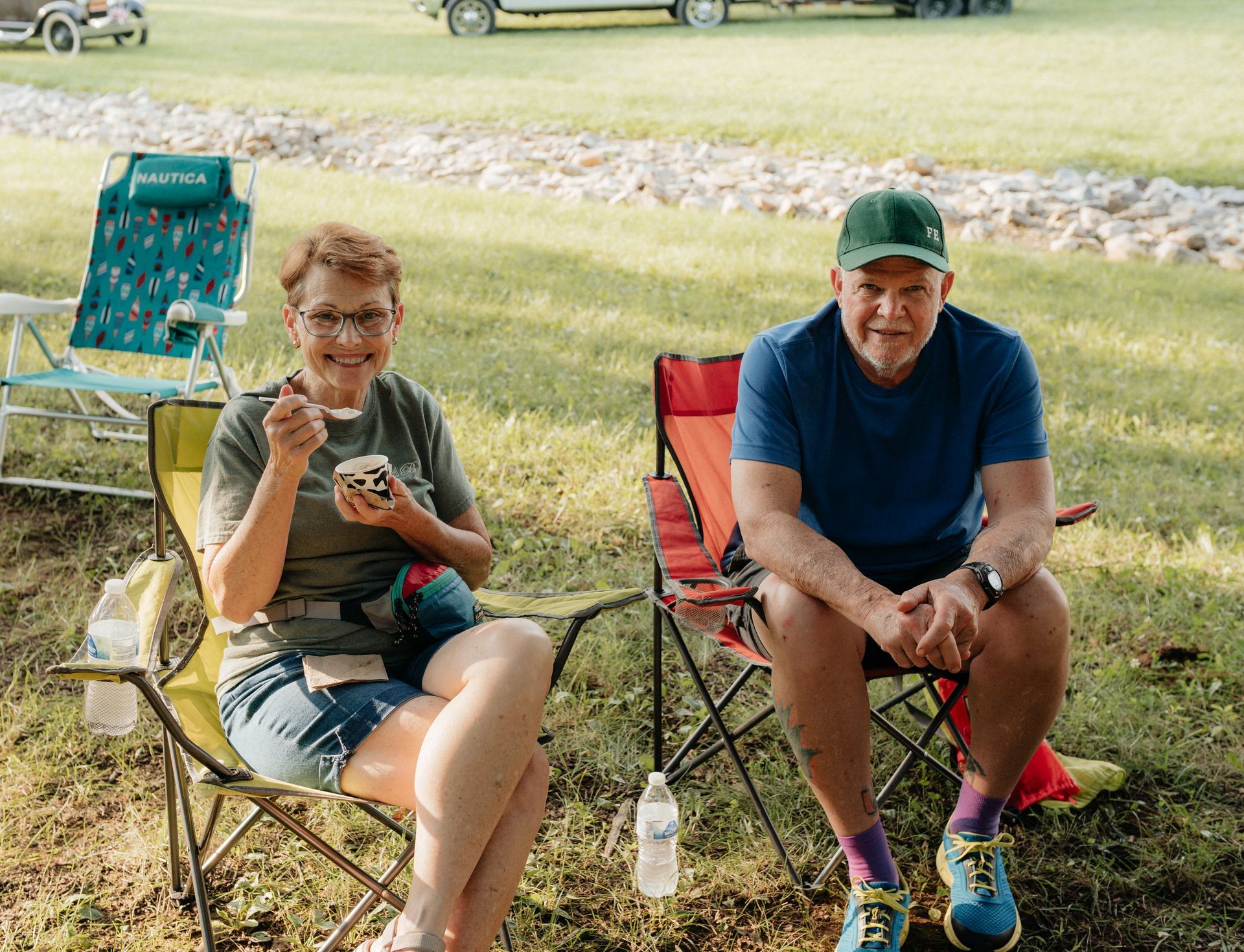 Two people sitting in chairs on grass, eating ice cream and wearing a hat.