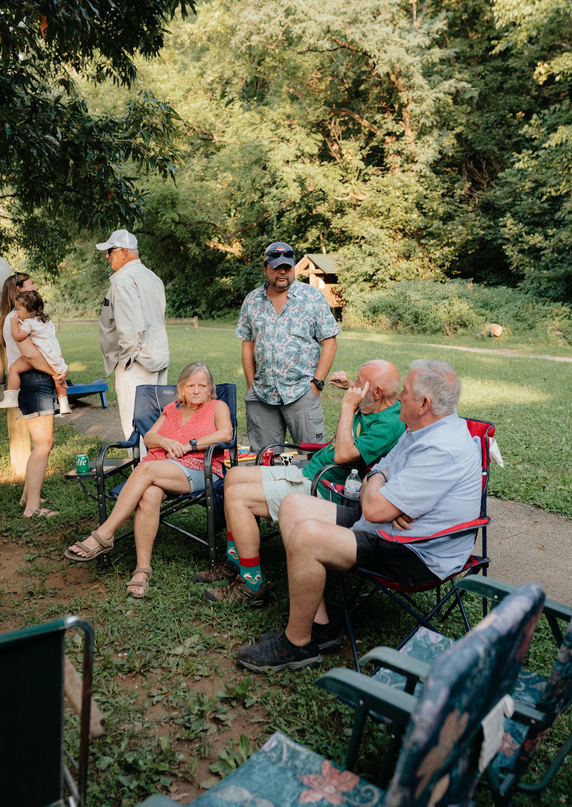 Group of people relaxing outdoors, sitting and standing, trees in the background, sunny day.