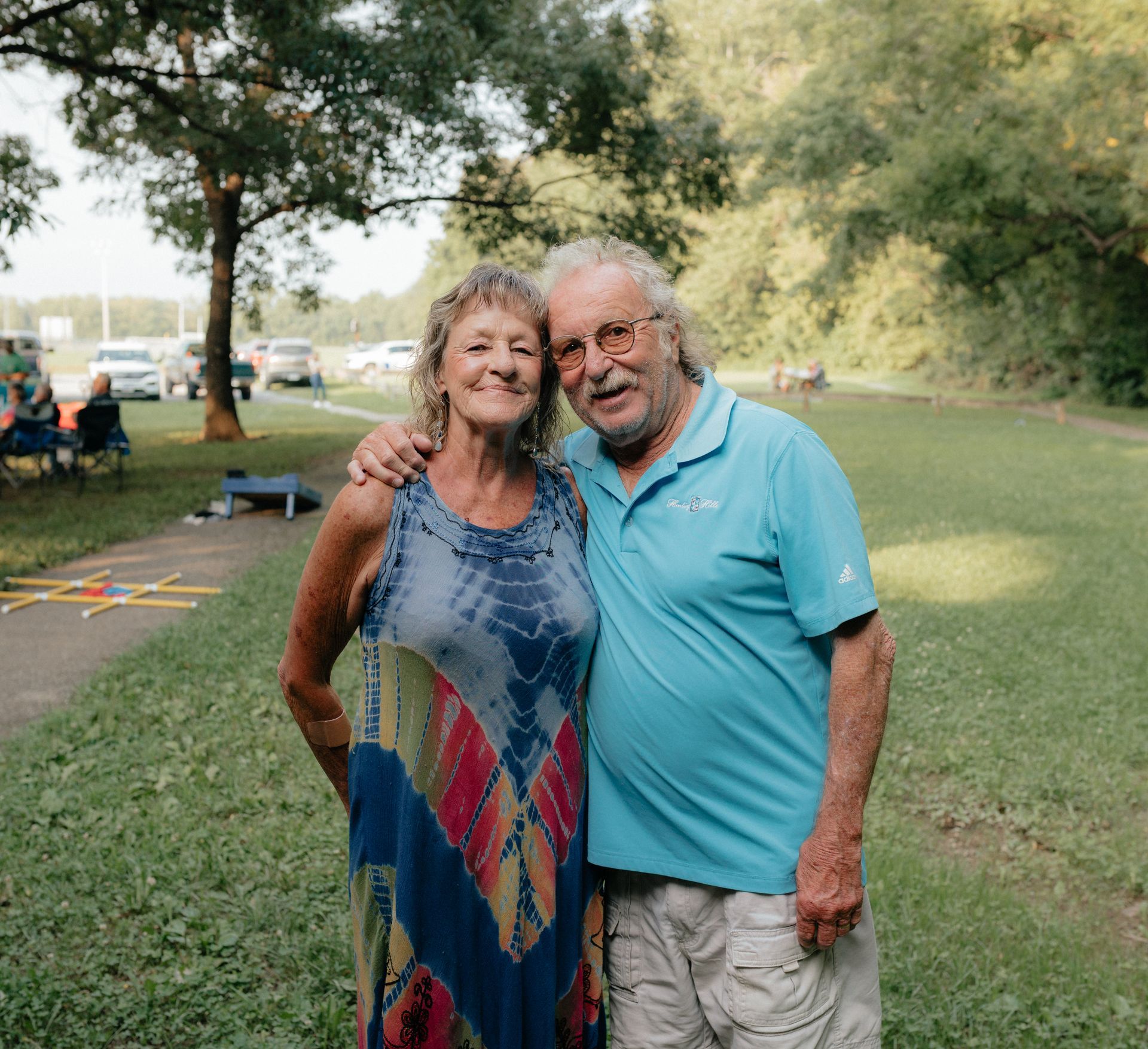 An older couple smiles, arm in arm, on a grassy lawn; woman wears a tie-dye dress, man in a blue polo shirt.