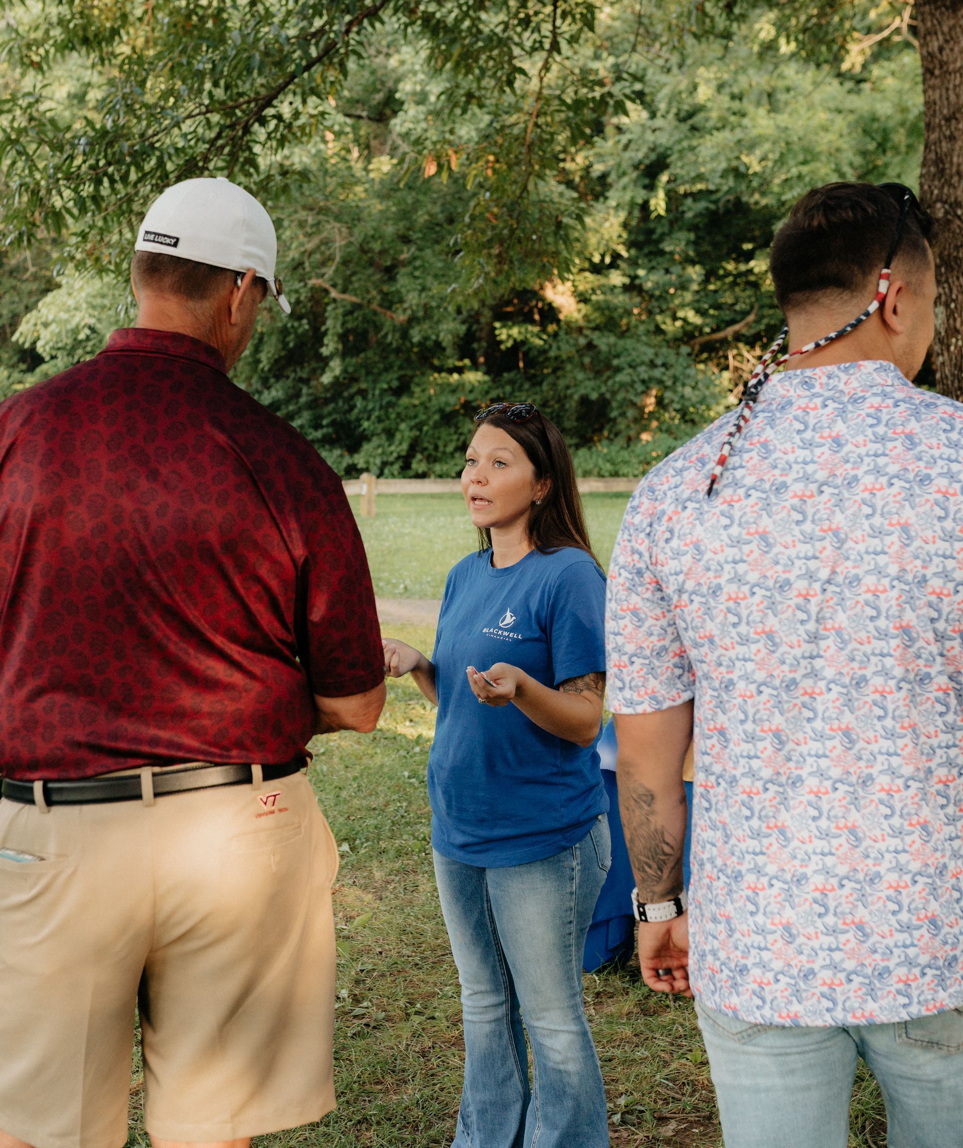 Three people talking outdoors. Woman in blue shirt gestures, two men face her; one in red, one in patterned shirt.