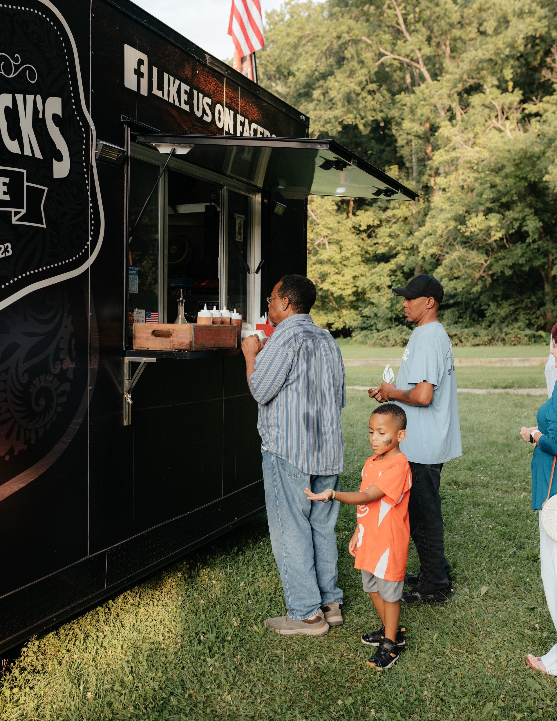People ordering from a black food truck with American flag. Man and child are closest to the truck.