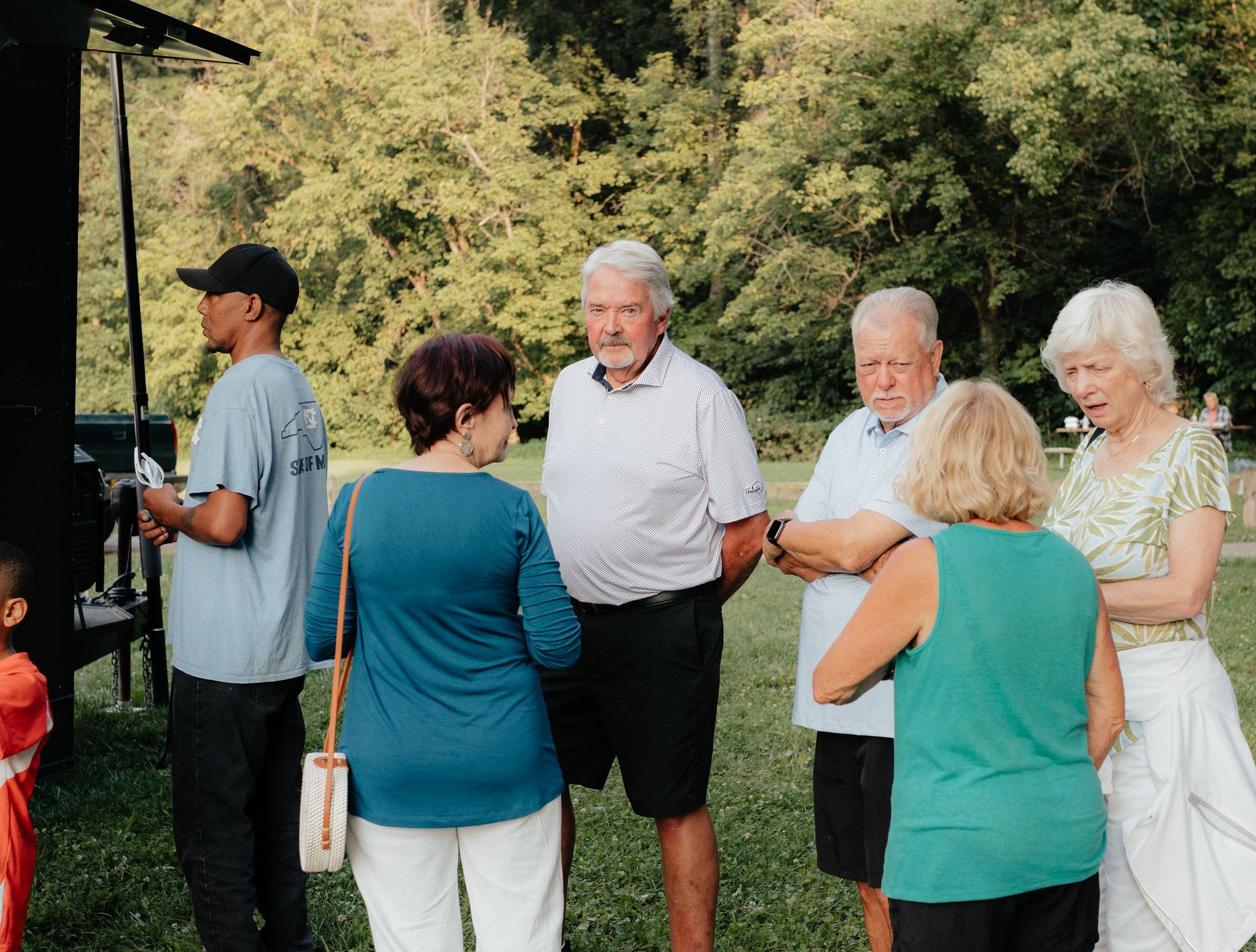 Group of people outdoors talking, including men and women; some are looking toward a dark object. Trees and greenery in the background.