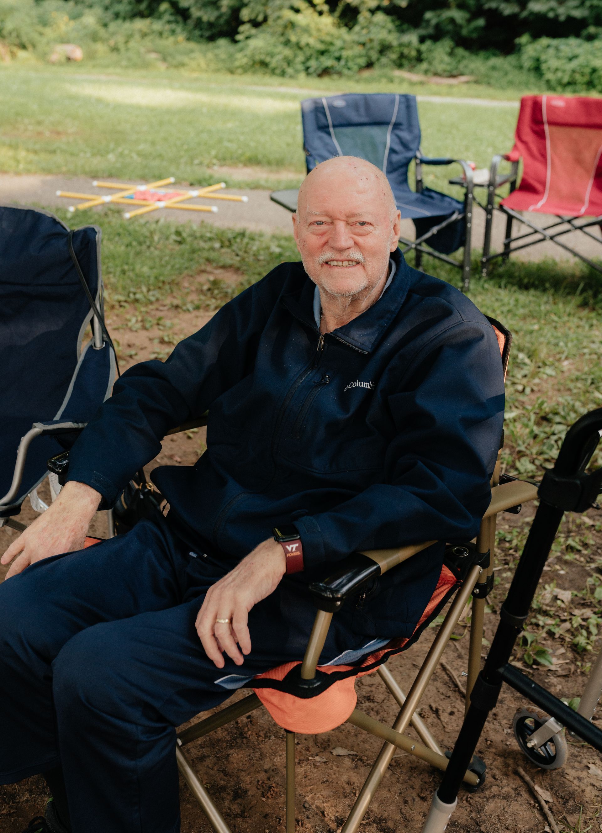 Older man with bald head smiles, seated outdoors in folding chair. Wearing blue jacket and pants.