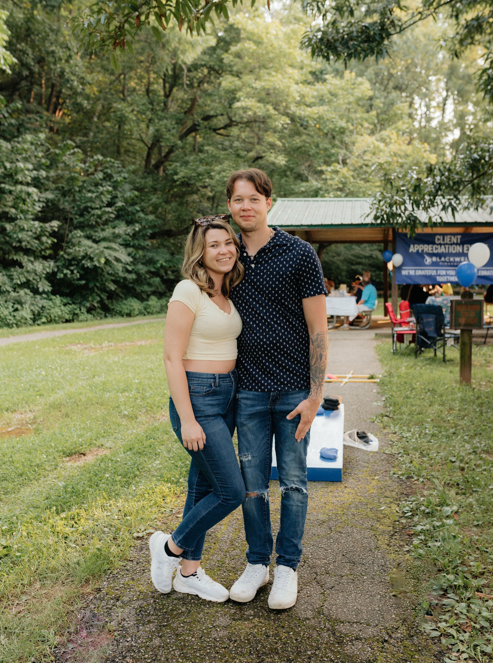 Couple posing outdoors. Woman in yellow top, jeans, white shoes. Man in blue, patterned shirt, ripped jeans, white shoes.
