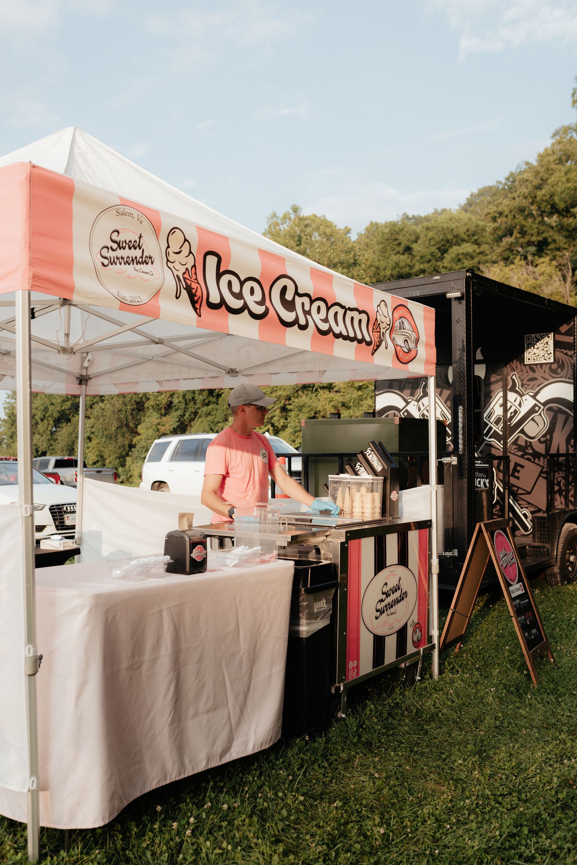 Ice cream stand with a person serving, white and pink canopy. Outdoors, sunny day.