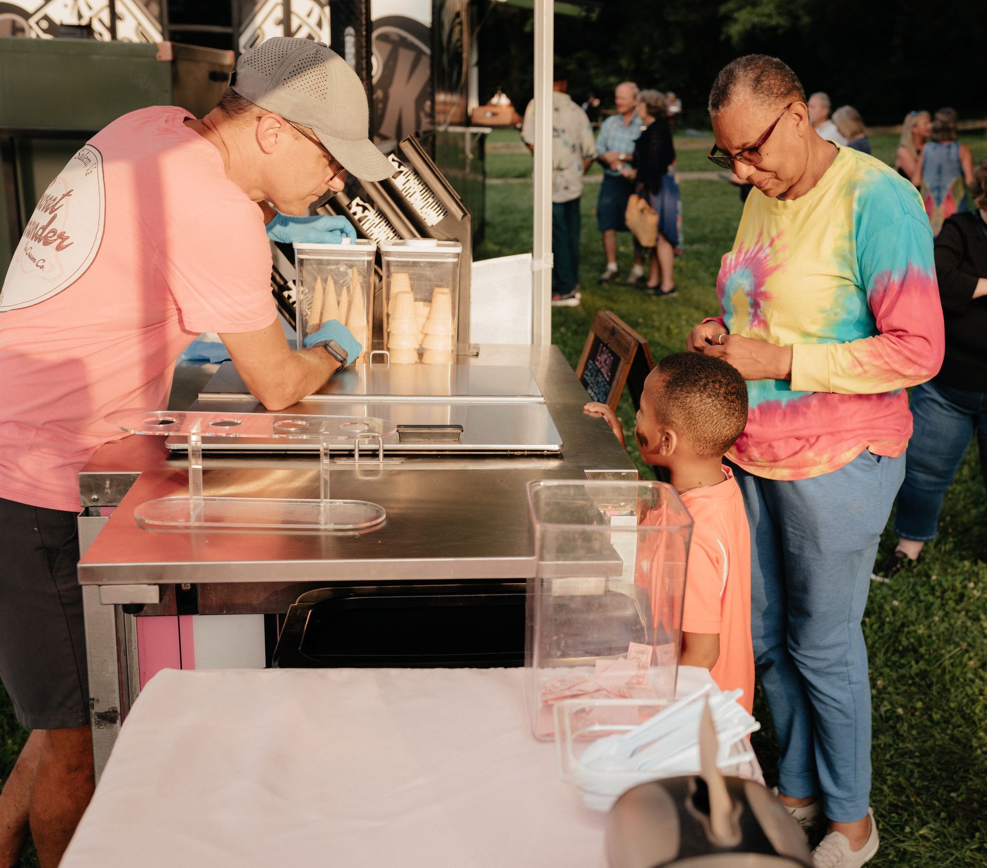 Person serving ice cream to a child and a woman at an outdoor food stand; people in the background.