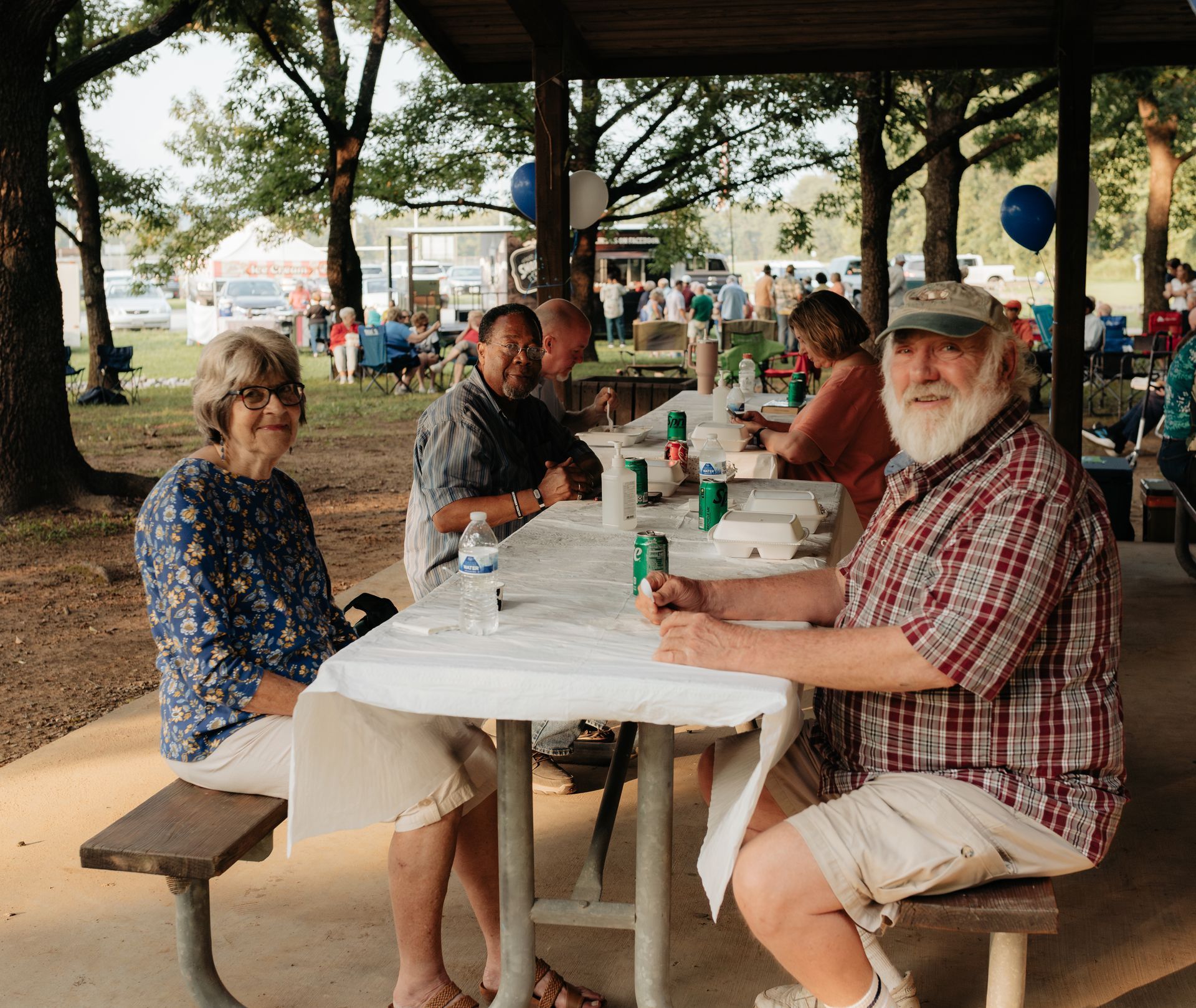 Group of people at a picnic under a shelter. Elderly man smiles, others at tables with food and drinks. Outdoors, trees.