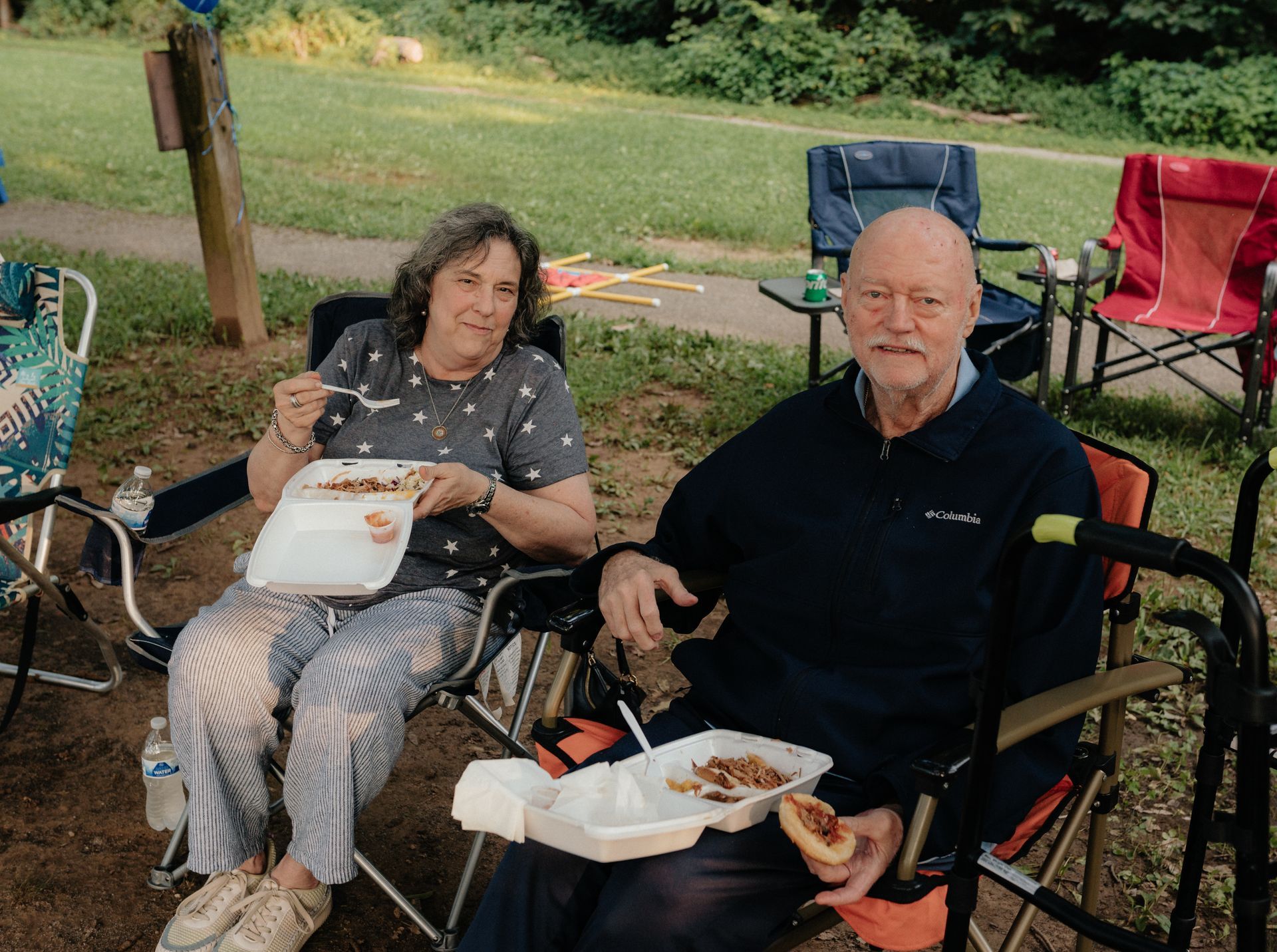 Elderly couple eating outdoors, sitting in camping chairs. Woman eating, man with a walker. Park setting.