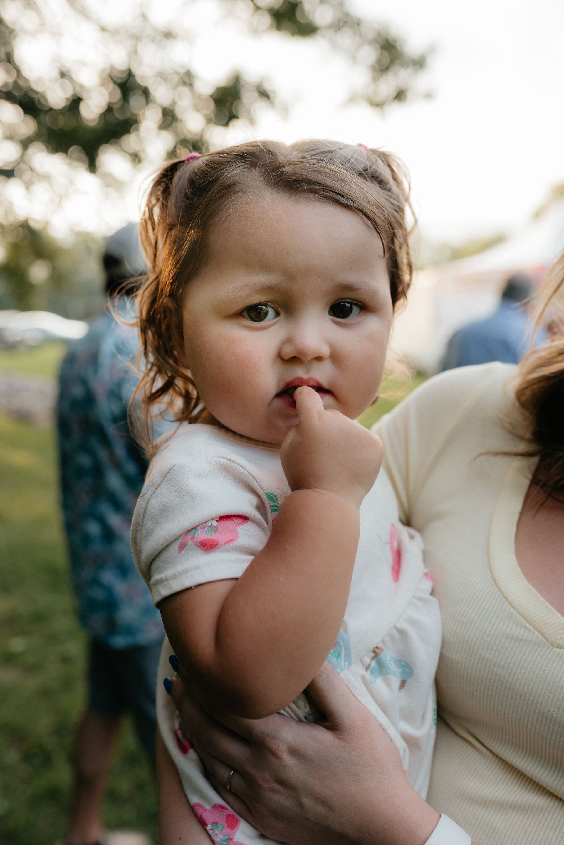 Toddler held in arms, looking at the camera with finger in mouth, outdoors in sunlight.