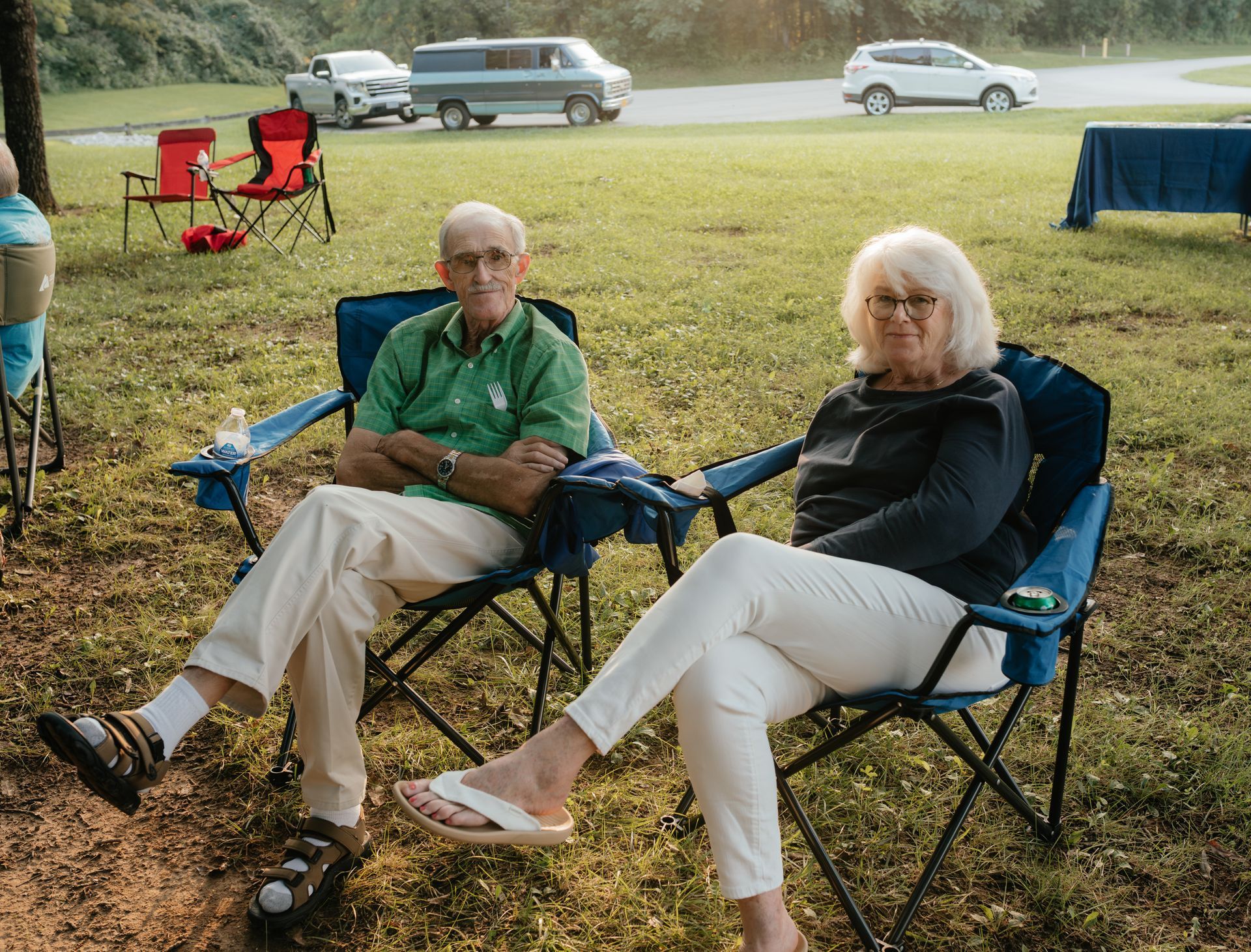 An elderly couple sits in camp chairs, looking at the camera. They are in a grassy area, with cars in the background.