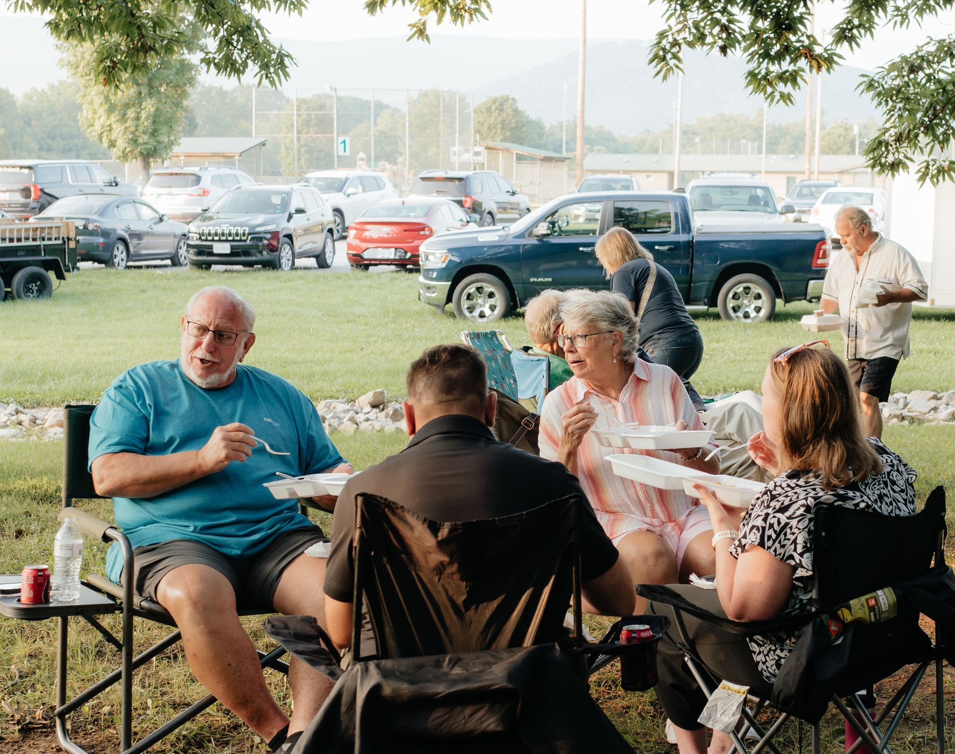 Group of people eating at a picnic, outdoors near cars.