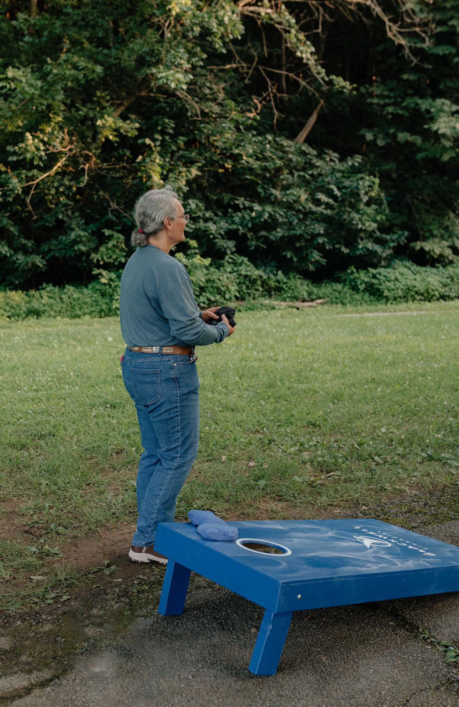 Woman in jeans throws a beanbag at a blue cornhole board in a park.