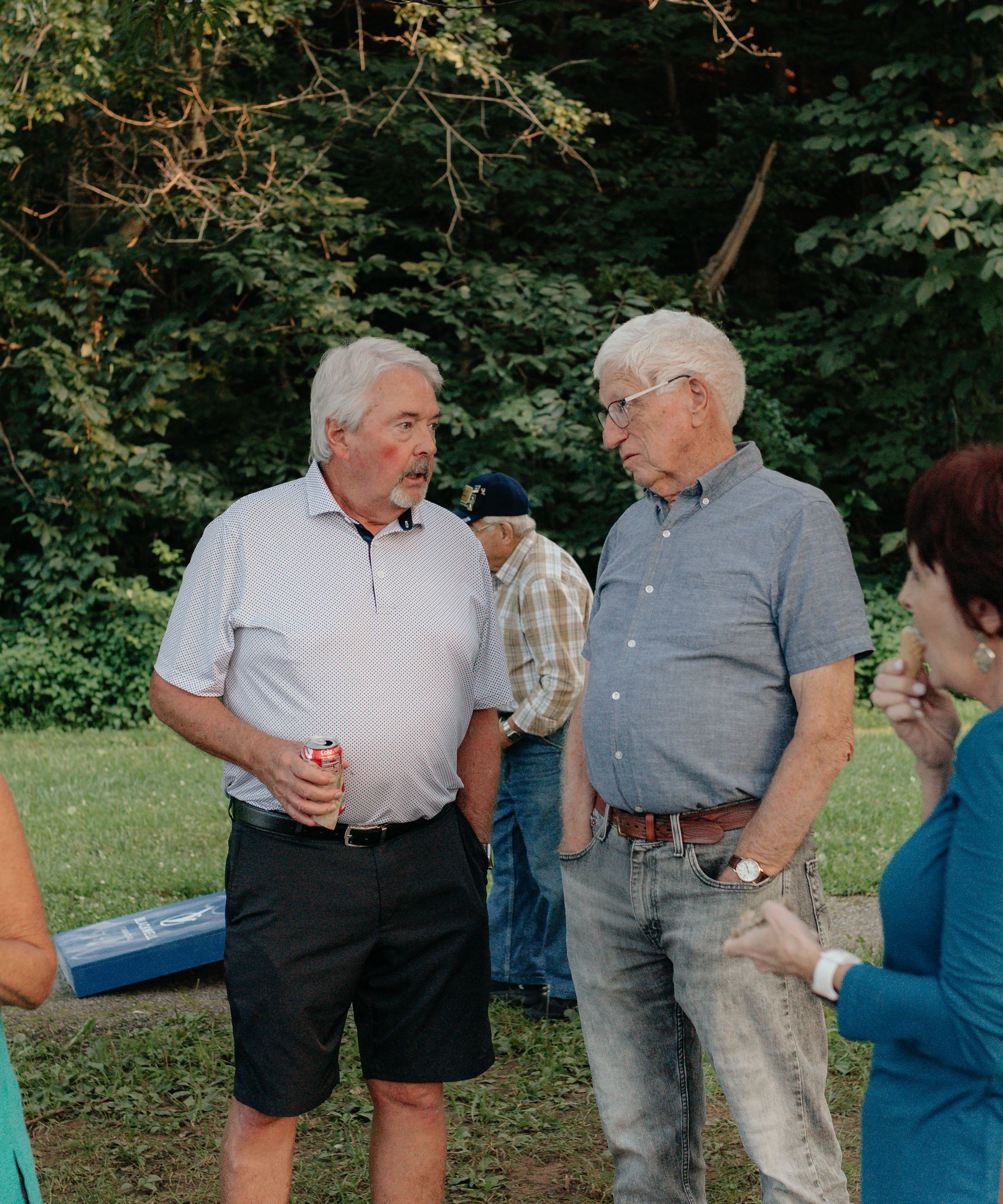 Two older men conversing outdoors; one in shorts, the other in jeans, and a woman in blue nearby.