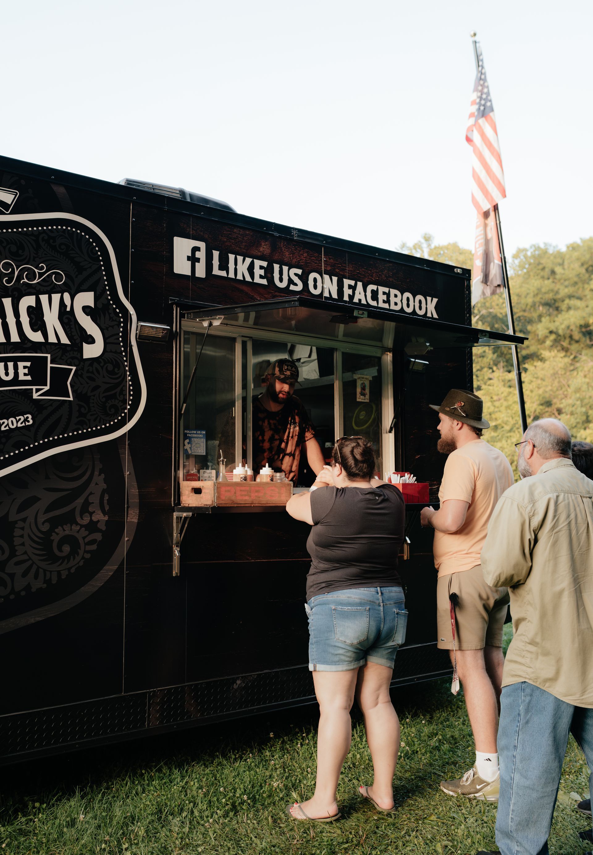 People ordering food from a BBQ food truck with a US flag.