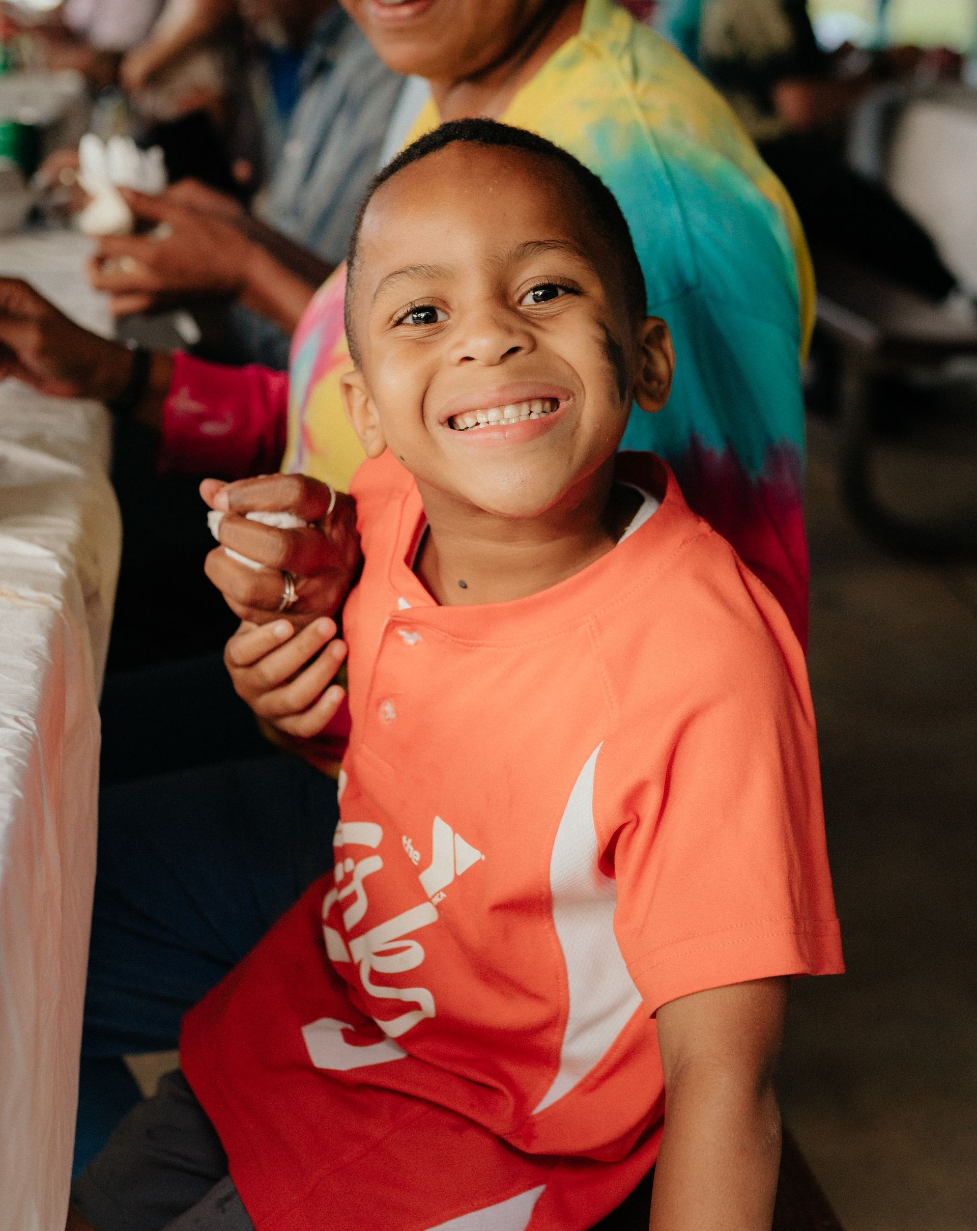 Smiling boy in an orange shirt with a white design; seated with an adult at a table.