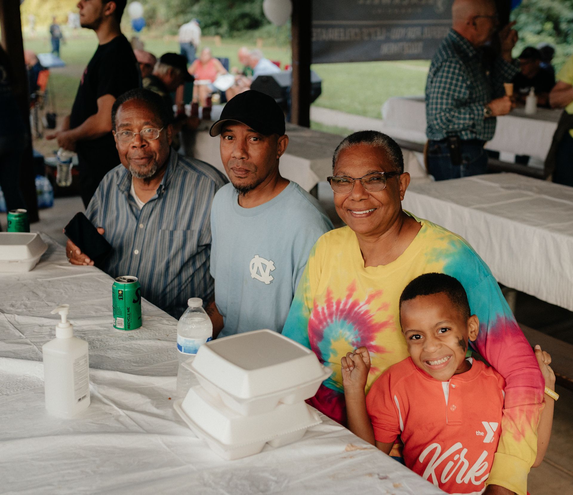 Family smiling at a table during an outdoor event. Includes an older man, son, daughter, and young child with a painted face.