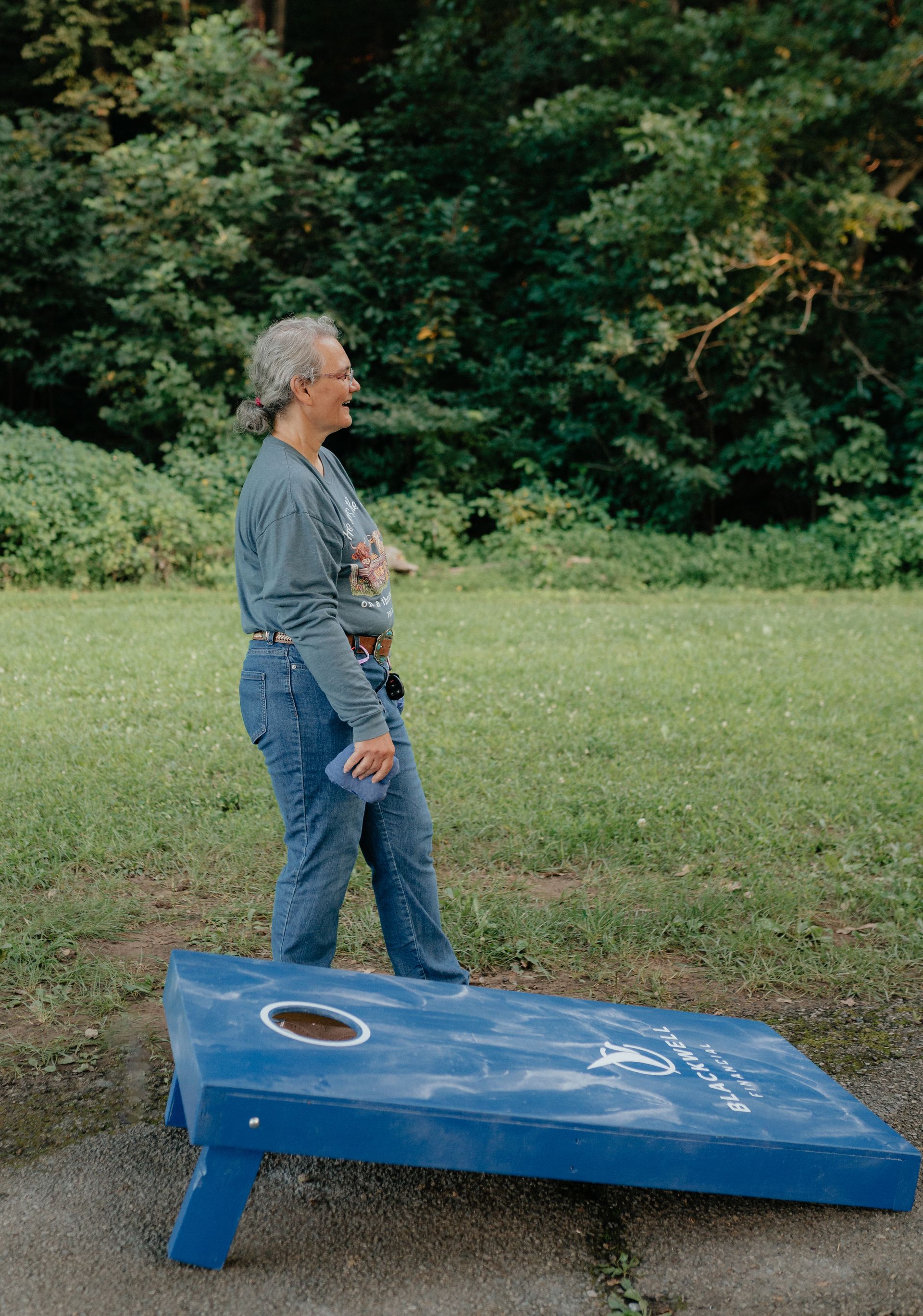 Woman playing cornhole in a park. Blue cornhole board, green grass, trees in background. She's smiling.
