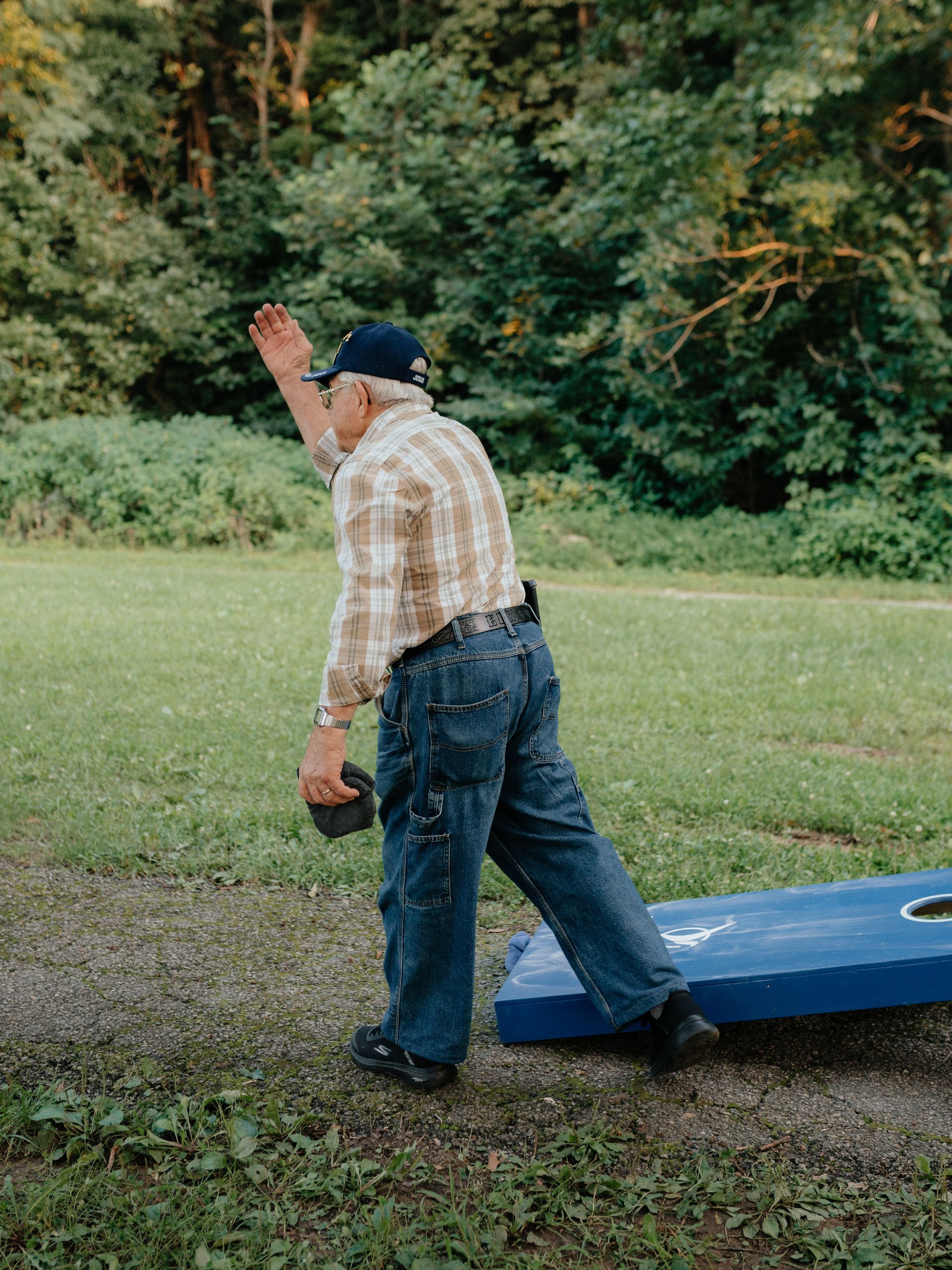 Man in plaid shirt and jeans throwing a bean bag at a cornhole board outdoors.