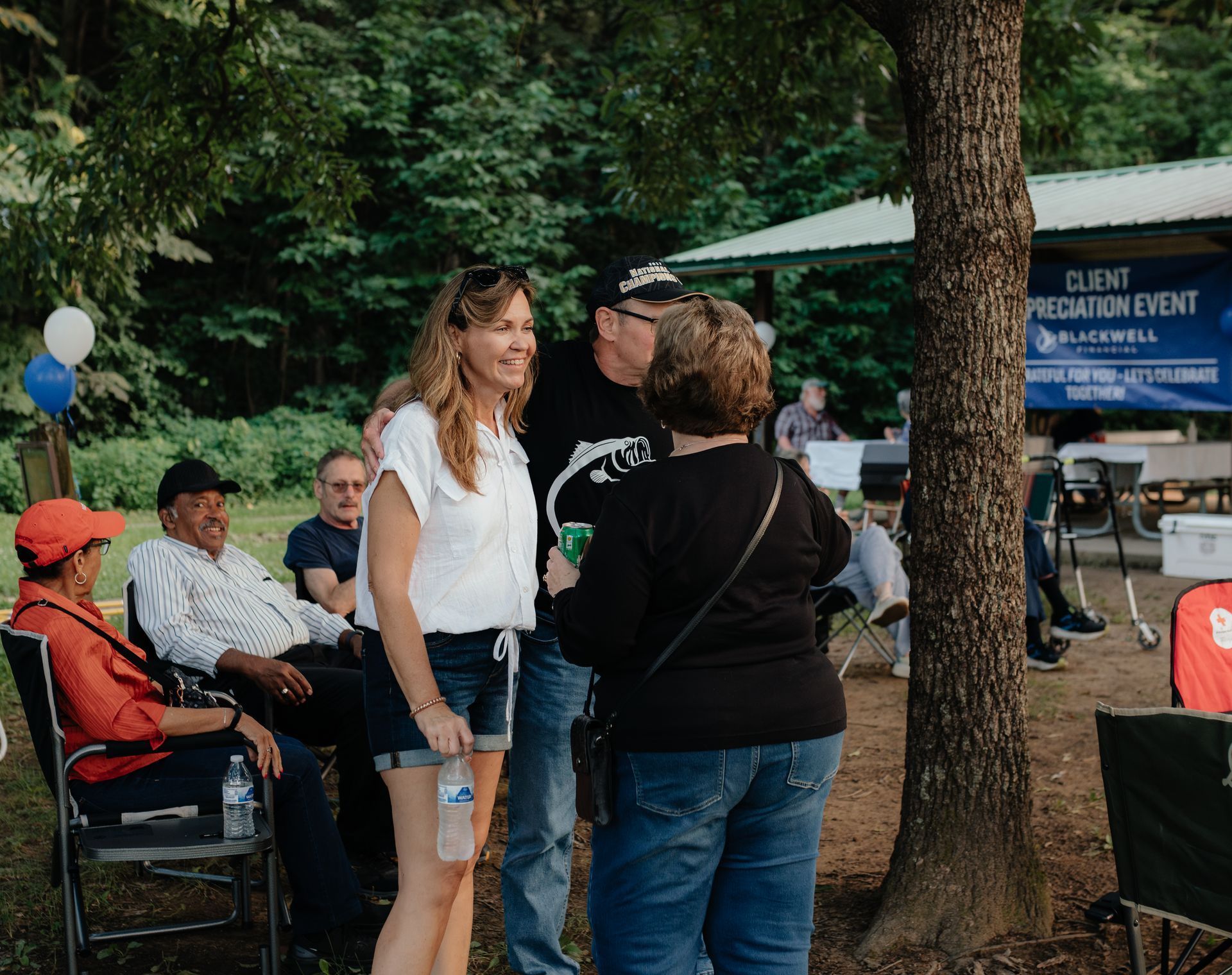 People at a client appreciation event outdoors, socializing. Woman smiles, talks with others, near a tree, chairs, and event sign.