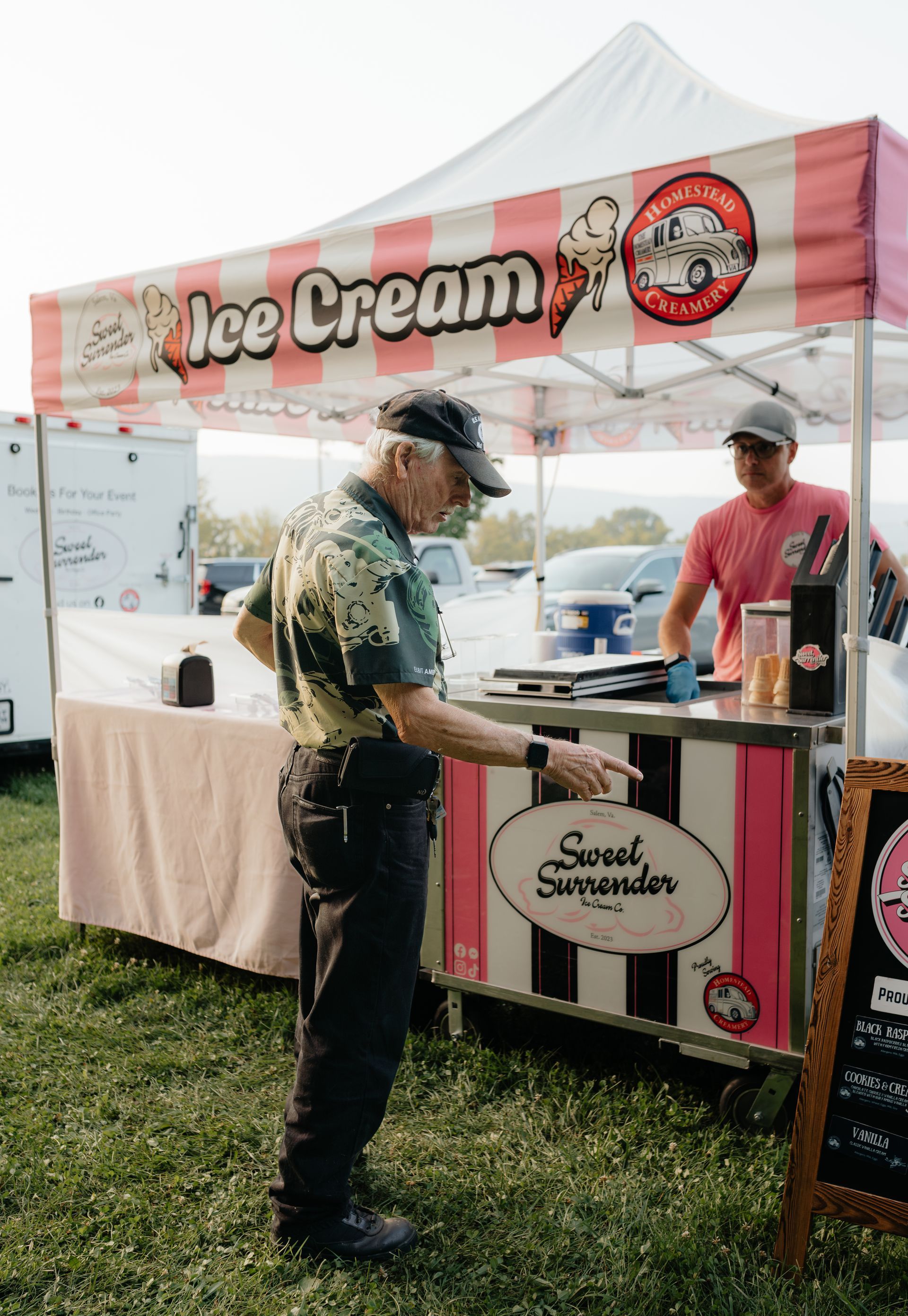 Older man ordering ice cream from a pink and white striped stand at an outdoor event.