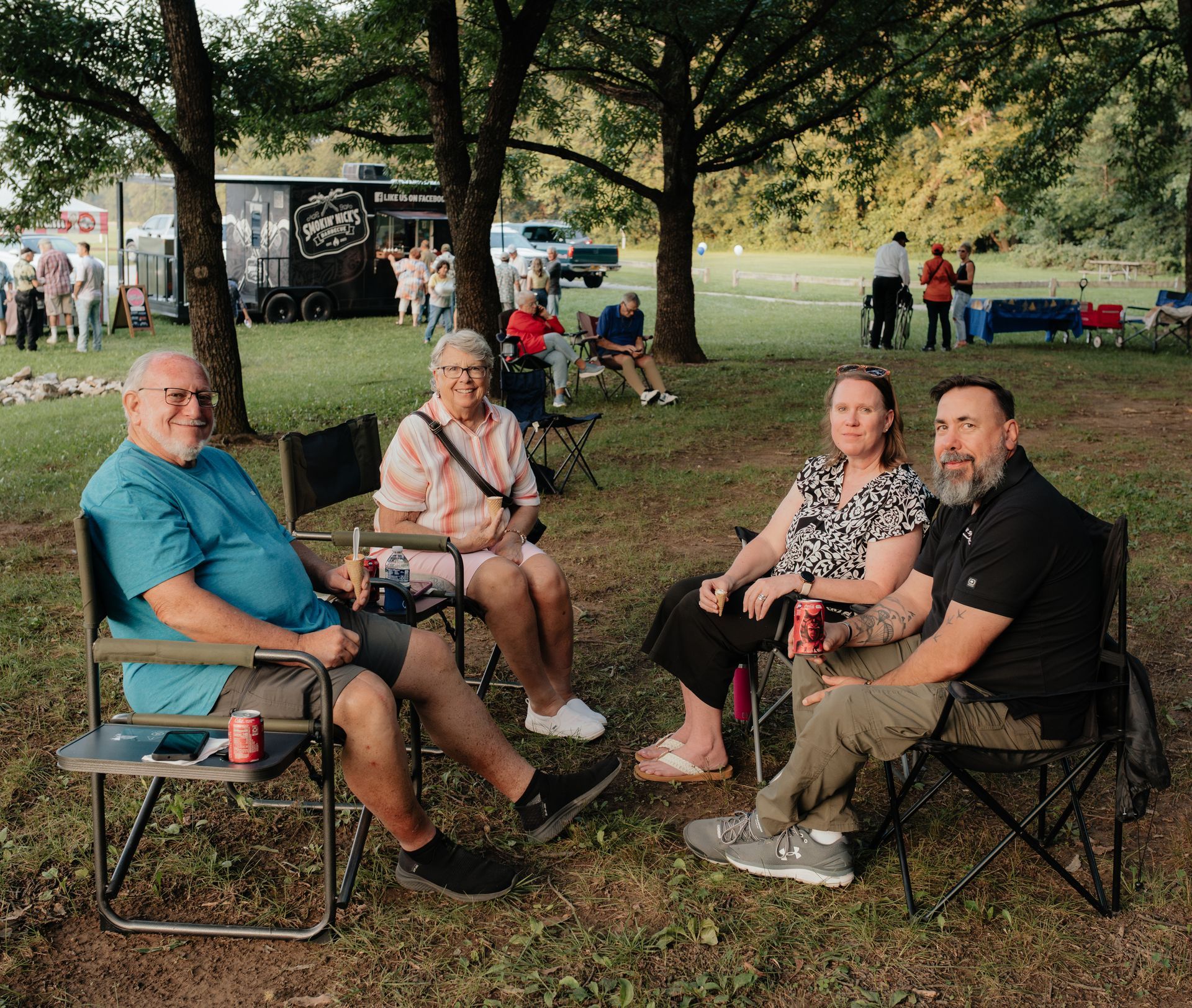 Four people seated in camping chairs enjoying an outdoor event. Others are visible in the background near a food truck.