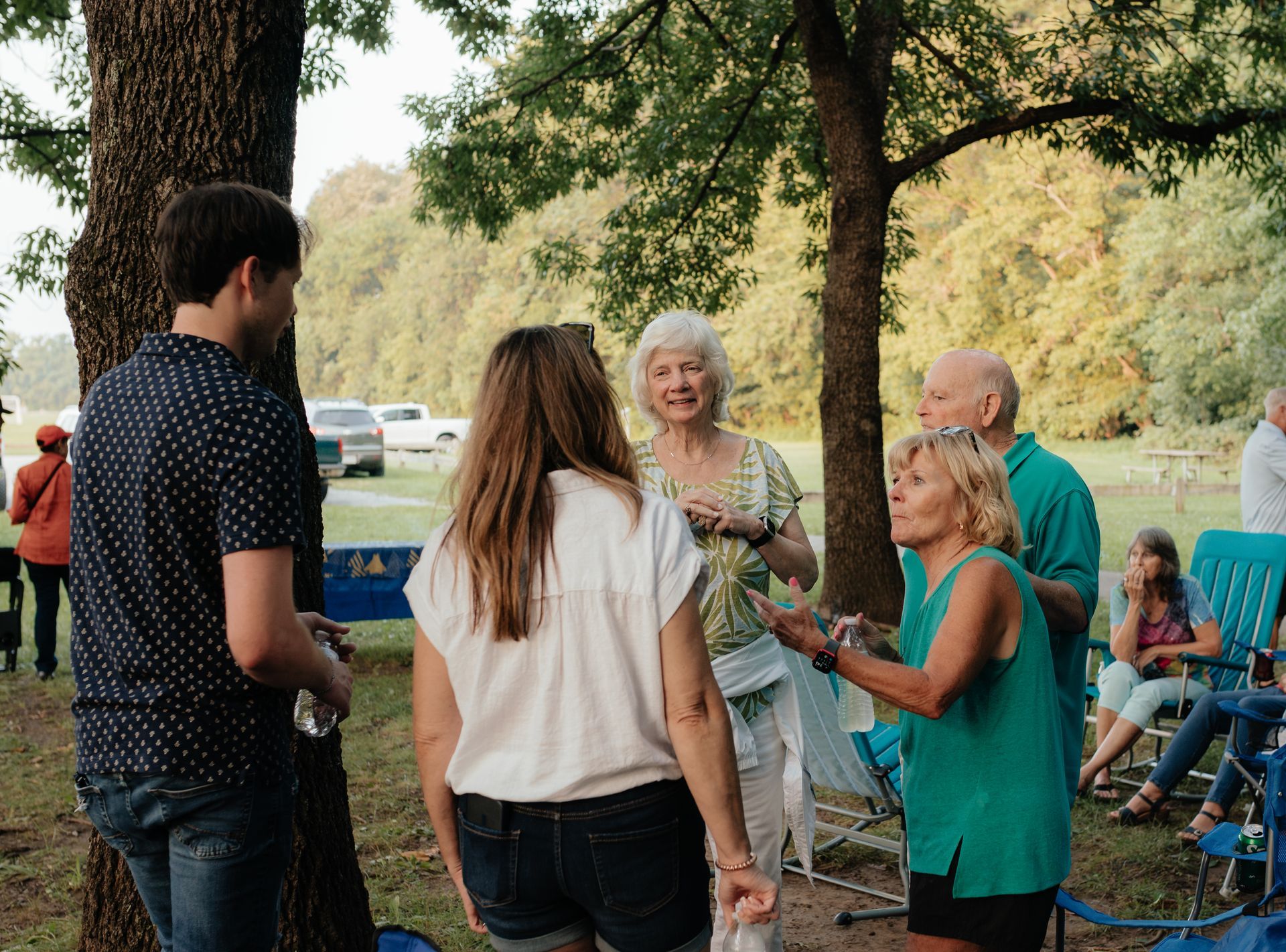 People socializing outdoors, under trees. A man in blue shirt talks to a woman. Elderly couple nearby, other people in background.