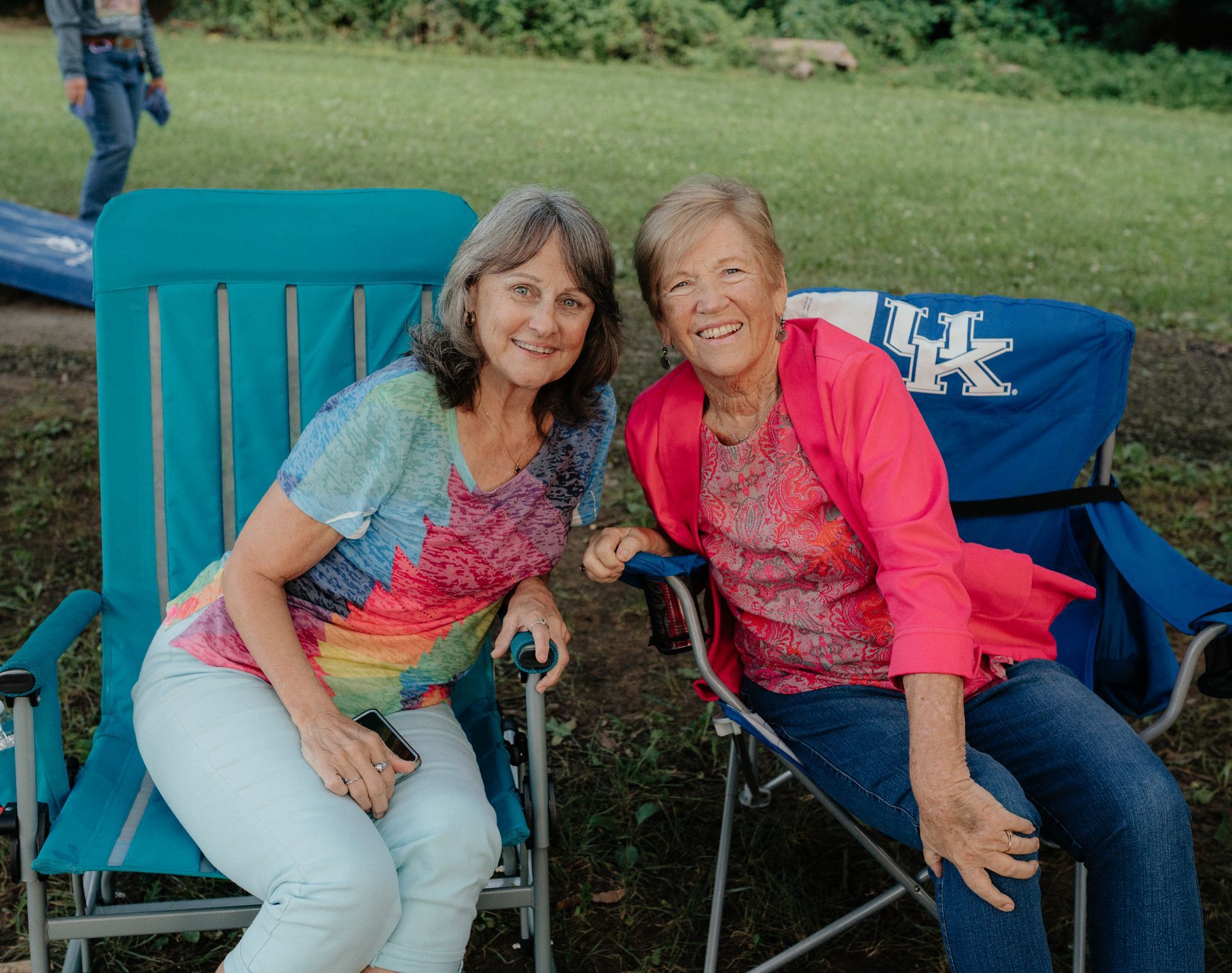 Two women smiling, sitting in folding chairs outdoors, one wearing a tie-dye shirt, the other a pink jacket.