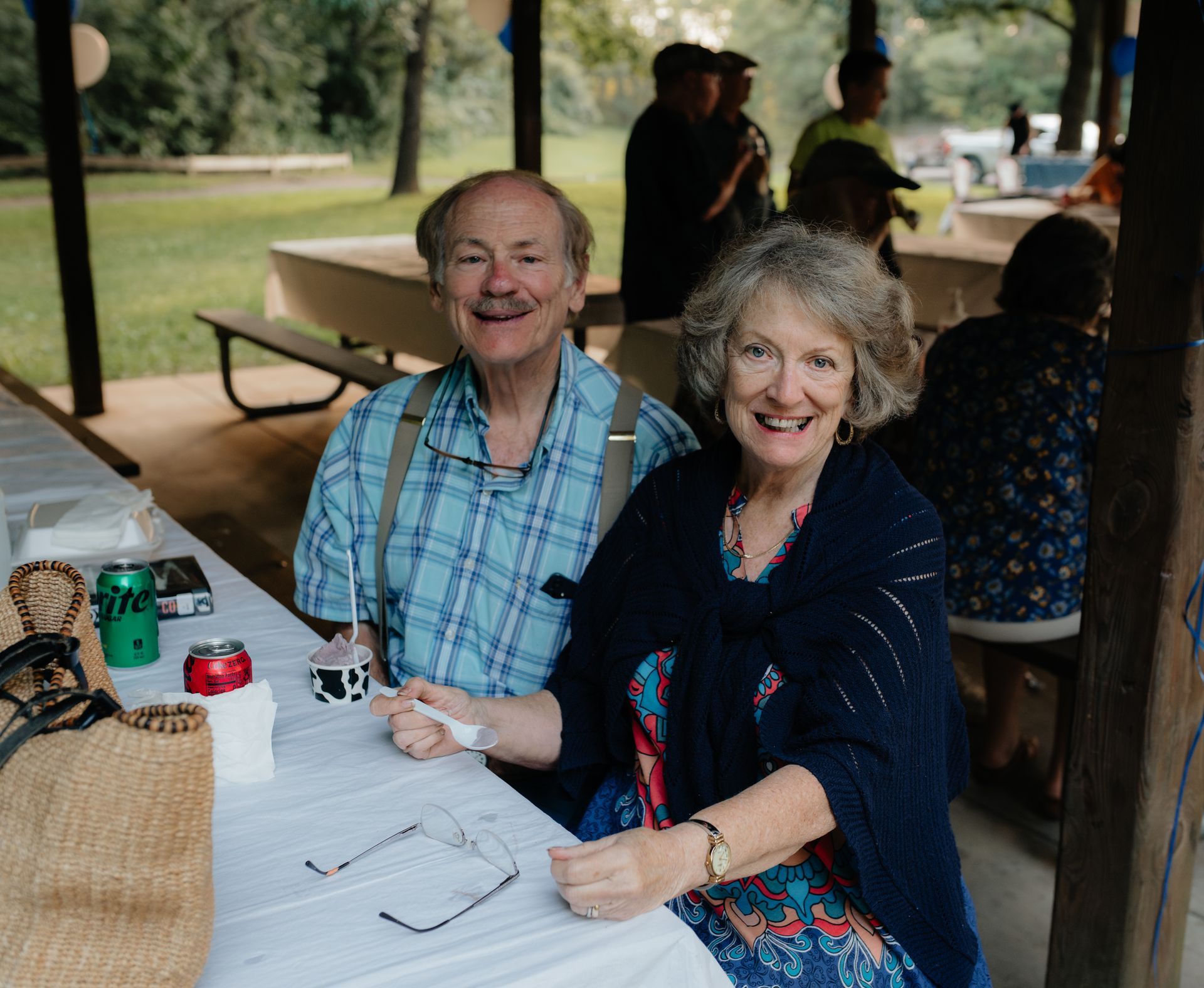 Smiling older couple at a picnic table, outdoors. Man in blue plaid shirt with suspenders; woman in blue floral dress.