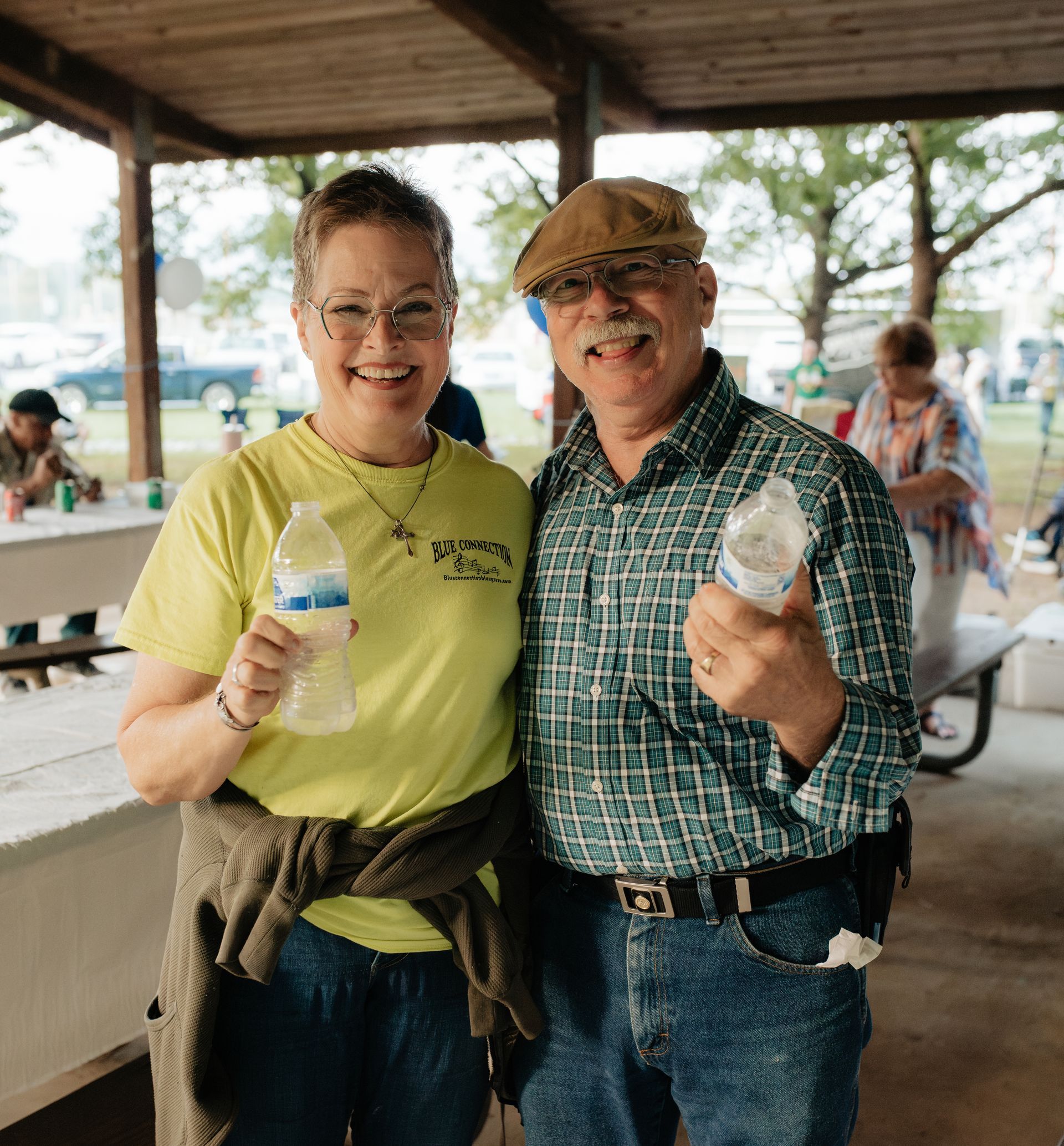 Two people smiling, holding refreshments, at an outdoor event under a shelter.