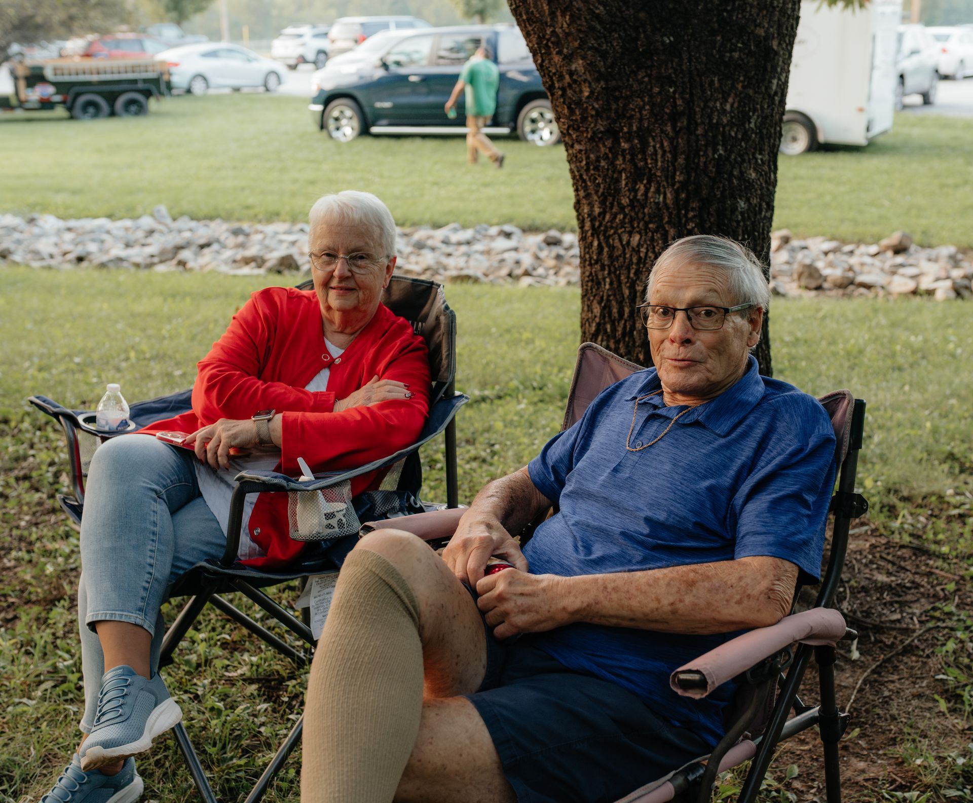 Two older adults sitting in folding chairs outdoors, a woman in red and a man in blue, with cars and a tree in the background.
