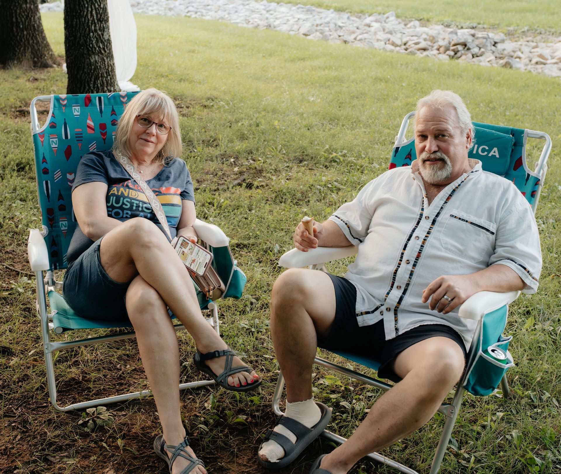Woman and man sitting in lawn chairs, grassy area. She wears glasses, he has a beard, both relaxed.