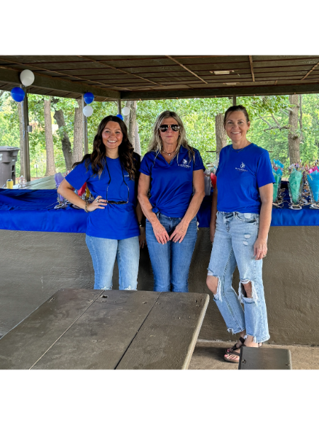 Three women in blue shirts are standing next to each other in front of a picnic table.