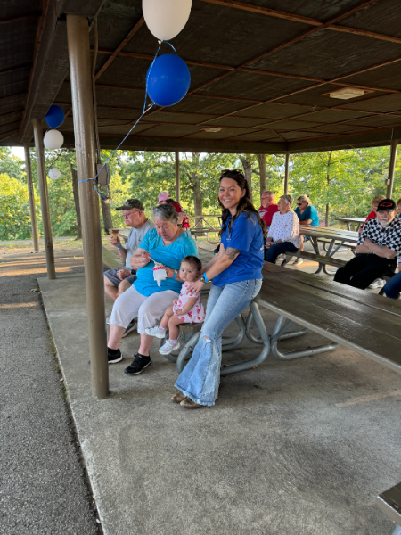 A group of people are sitting at picnic tables under a covered area with balloons hanging from the ceiling.