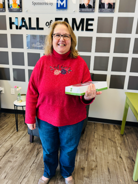 A woman in a red sweater is holding a box in front of a wall that says hall of fame