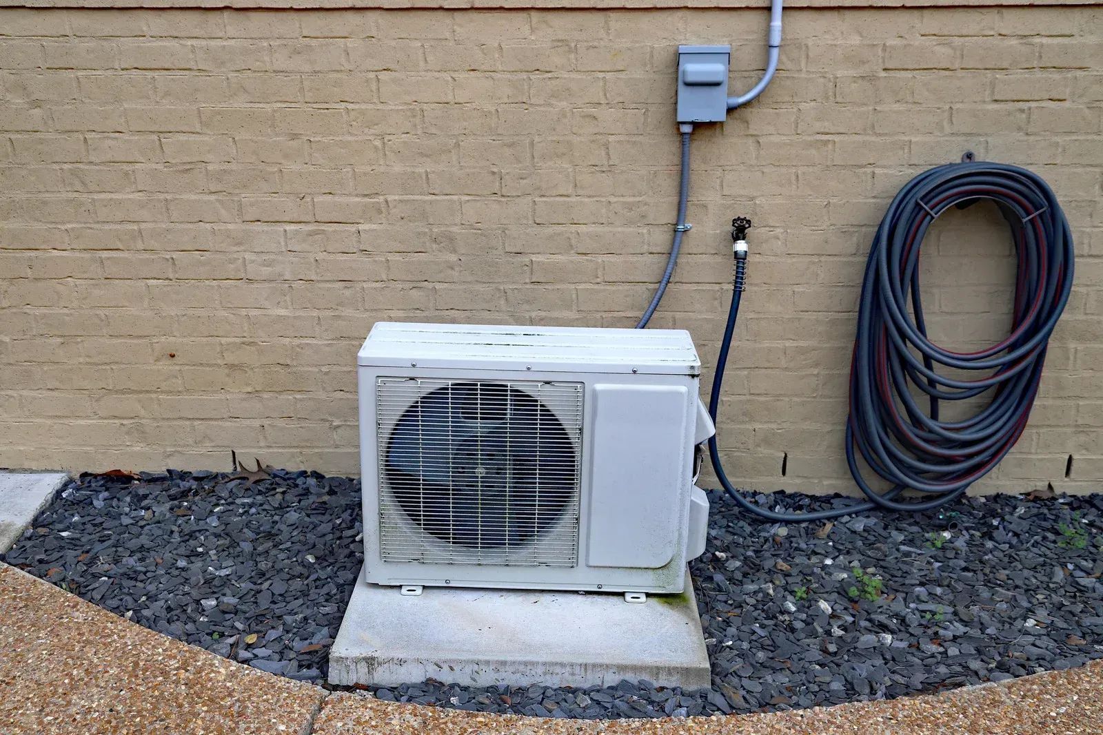 Air conditioning unit on a concrete pad next to a tan brick wall. A coiled hose is also present.