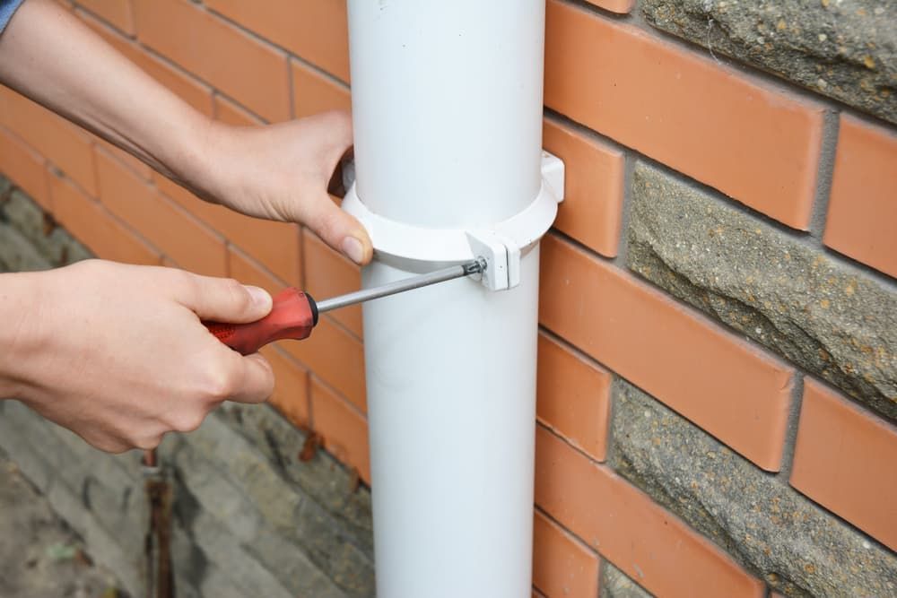 A Person Is Fixing a Drainpipe on A Brick Wall with A Screwdriver — Blair Merlehan Plumbing in Wagga Wagga, NSW