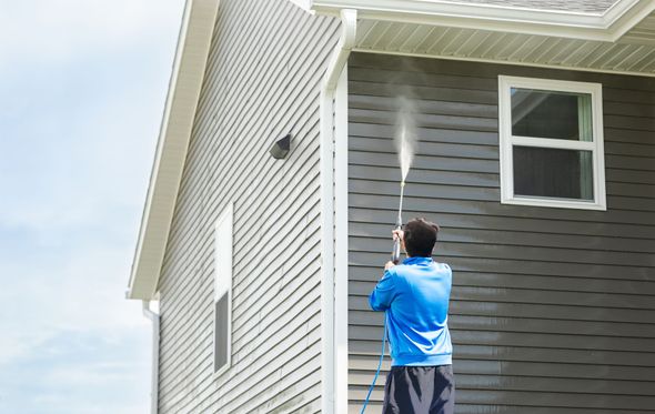 Person pressure washing the gray siding of a house on a sunny day.