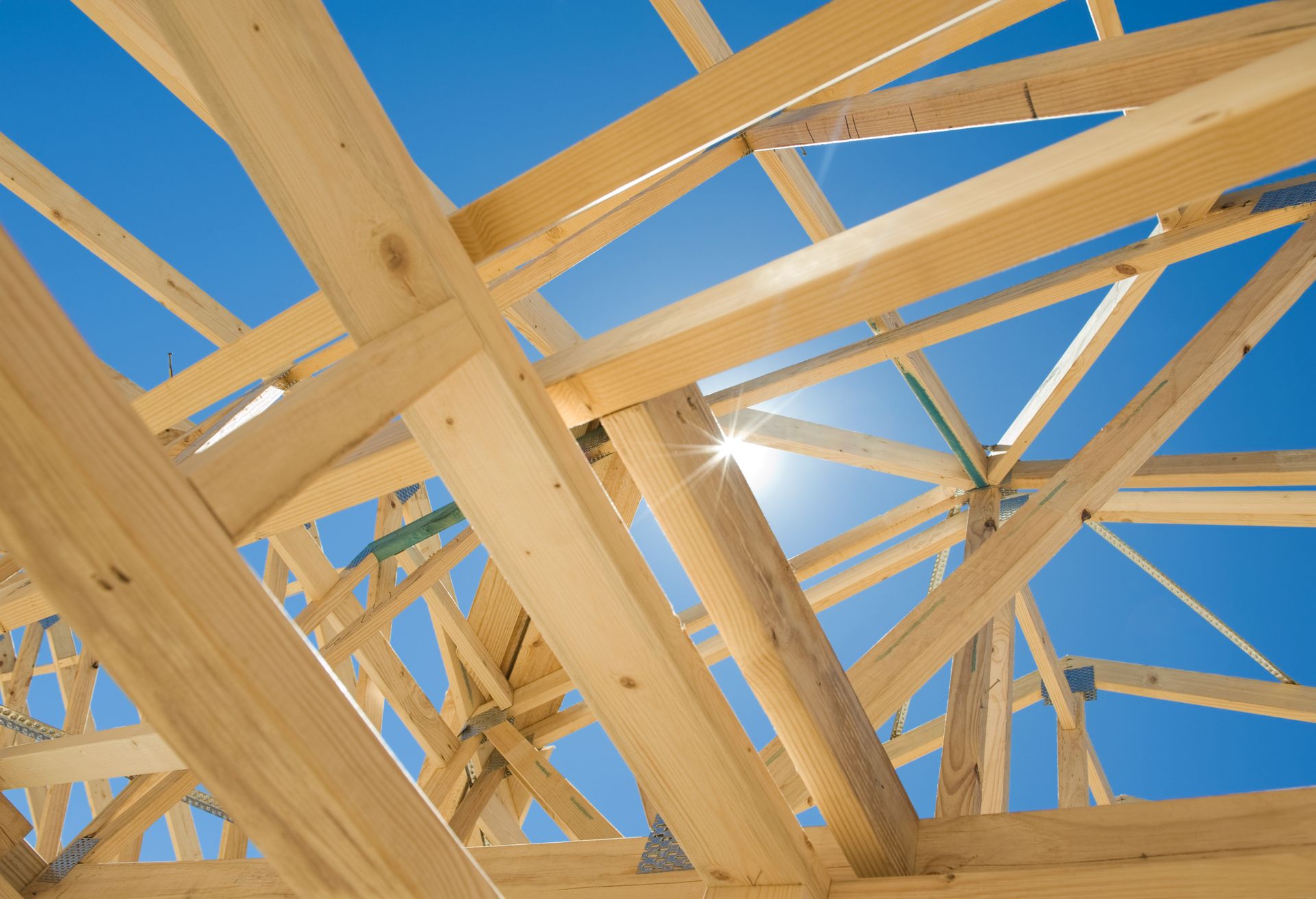 Wooden roof framing with intersecting timber beams viewed from below against a clear blue sky.