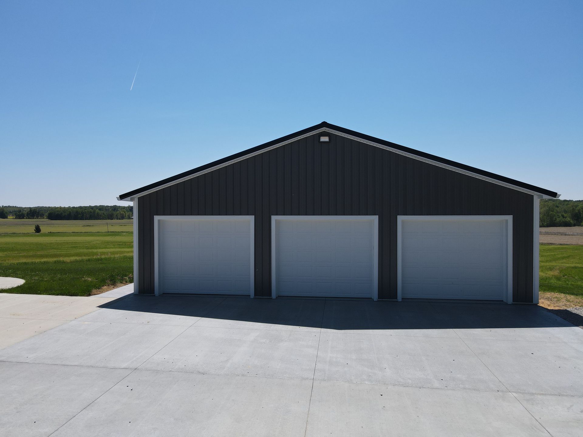 Dark metal garage with three closed white doors on a concrete apron in a grassy field